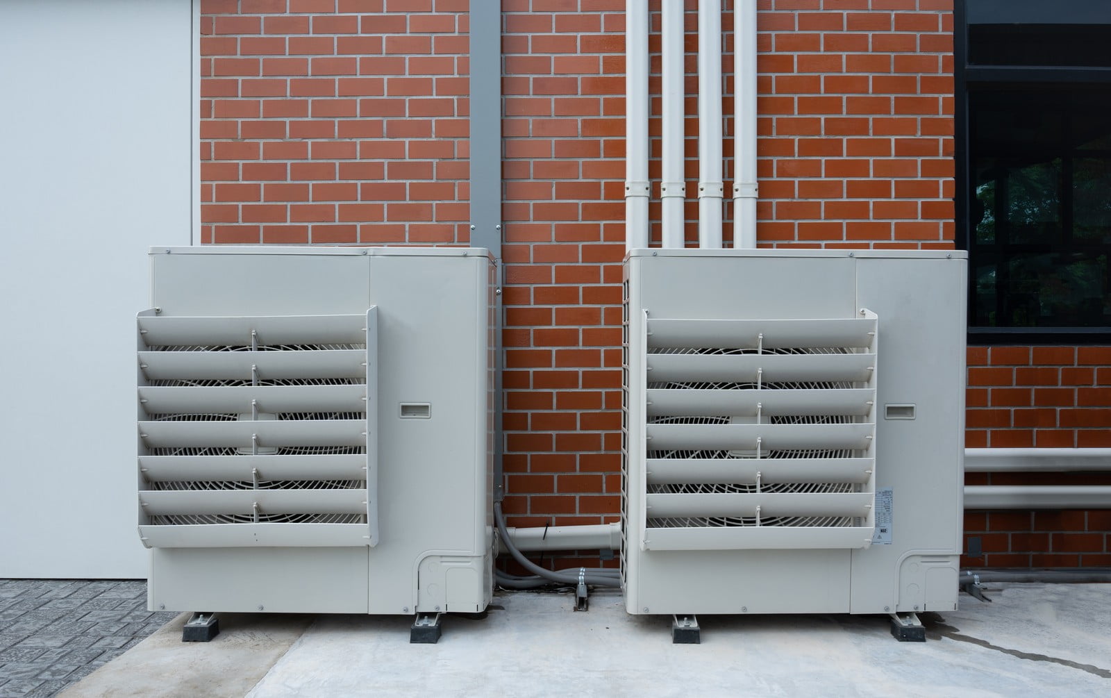 This image shows two large commercial or industrial air conditioning units placed outside a building. The units, consisting of metal enclosures with ventilation grills, are typically used to cool and circulate air as part of an HVAC system. White PVC pipes, likely part of the system's ventilation or drainage, run up the brick wall behind them. The building has a section with brickwork and another with a light-coloured façade, and there's a window showing a reflective glass pane on the right. Below the units, puddles suggest that condensation or drainage water has collected there. The ground seems to be a paved area, often used for placing such equipment.