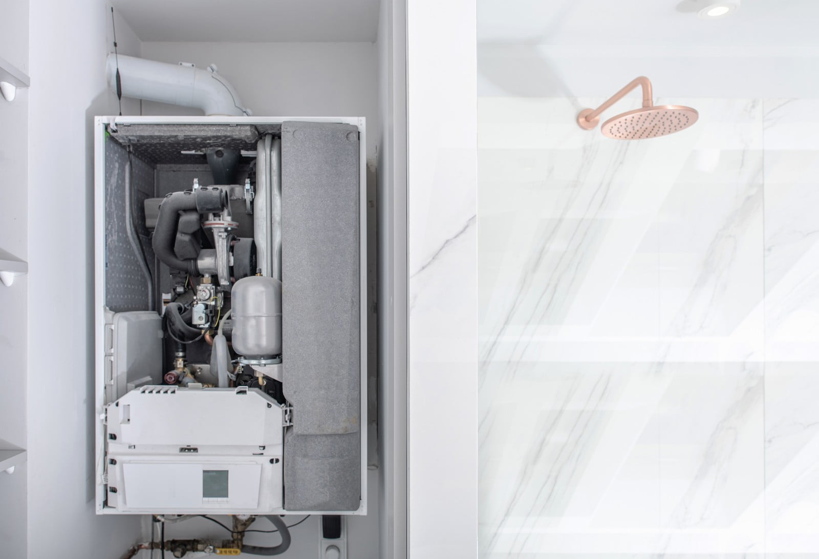 The image shows two distinct parts of a room. On the left, there is a domestic petrol-fired boiler with its front panel removed to reveal the internal components, such as the heat exchanger, pipes, and electronics that make up the central heating system. On the right, set against a wall with marble tiling, is a shower head fixed to the ceiling, indicating that this location is likely a bathroom or an area dedicated to showering. The contrast between the technical internals of heating equipment and the sleek, hygienic aspect of the shower space suggests a setting where utility meets comfort.