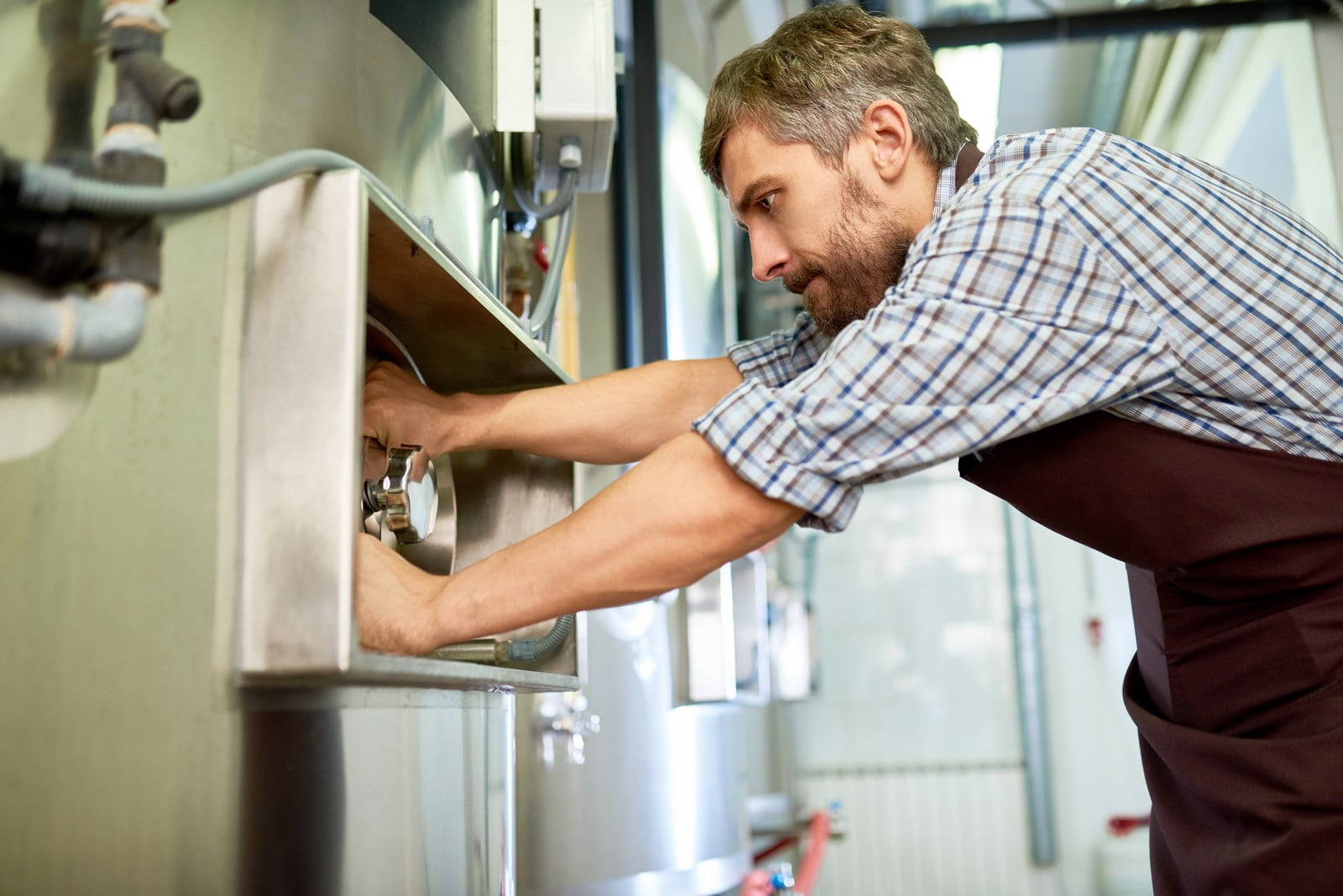 The image shows a man who appears to be working on some type of machinery or equipment. He is wearing a checkered shirt with rolled-up sleeves and brown trousers that suggest a work uniform. The man is focused on adjusting or fixing a component with his hands. It is not clear from the image what the exact nature of the work is, but it appears to be some sort of maintenance, repair, or inspection task. The equipment includes metallic parts and may be part of a larger system, possibly in an industrial or commercial setting. There are also some pipes and cables visible, which adds to the industrial atmosphere of the setting.