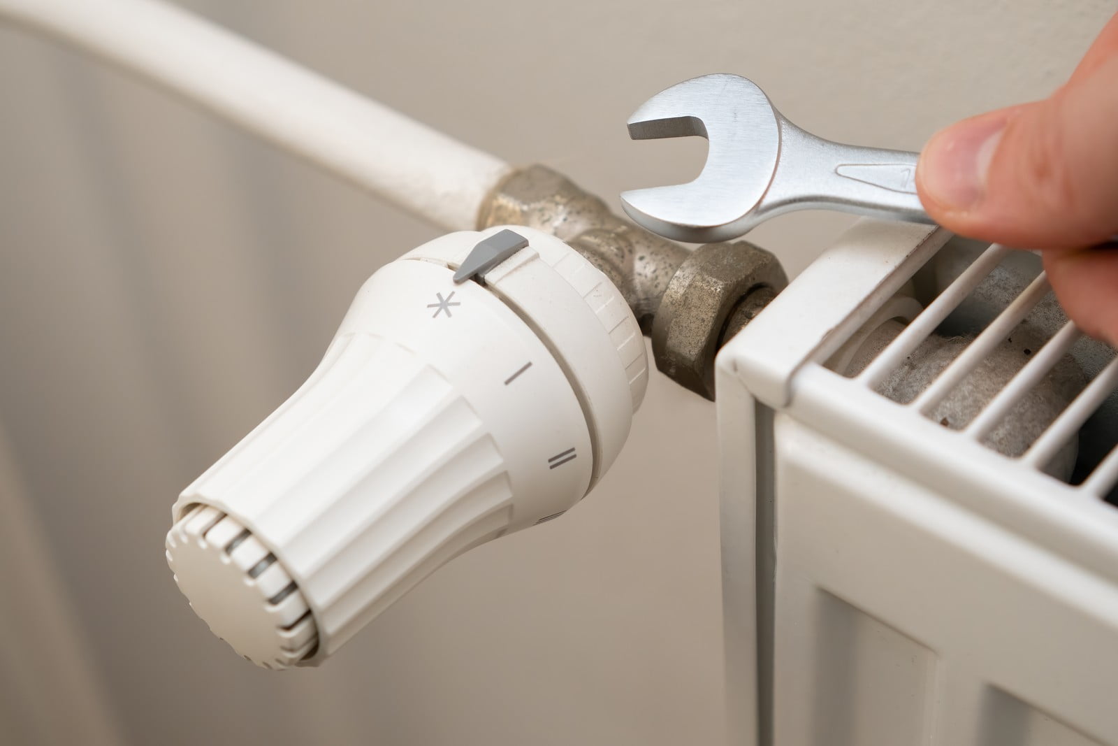 The image shows a person's hand using a spanner to adjust or work on a thermostatic radiator valve (TRV) which is attached to a radiator. TRVs are used to control the temperature of individual radiators and heating zones within a building. The radiator appears to be a compact style central heating radiator, which is commonly found in residential and commercial buildings. The setting on the TRV indicates a low temperature or possibly the 'off' position, represented by the star symbol. The overall scene suggests maintenance or adjustment of the heating system.