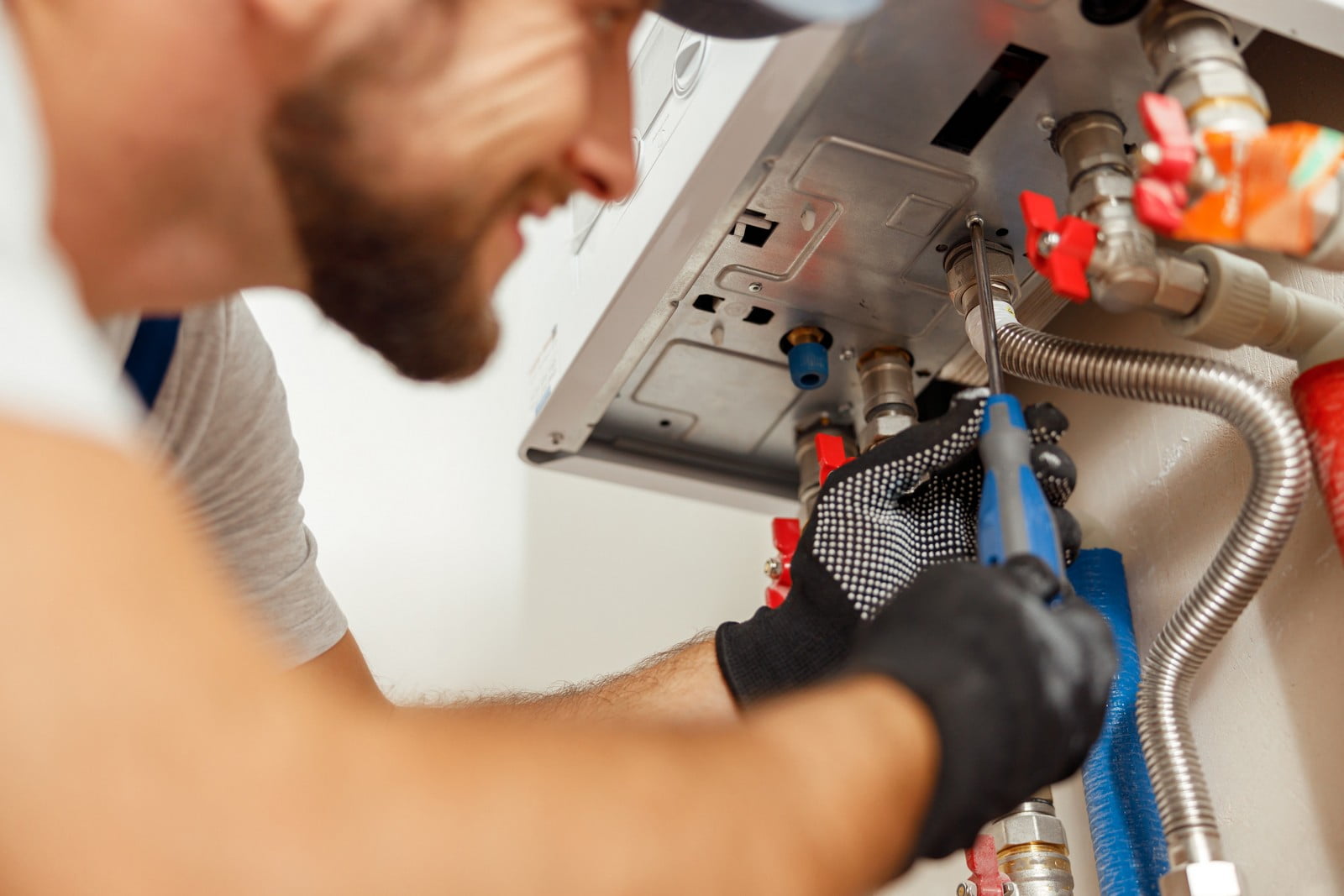 In this image, you see a person, likely a technician or plumber, working on a petrol boiler or water heater. The individual is wearing a short sleeve shirt and black gloves, suggesting an attention to safety or cleanliness. We see the boiler or heater open with visible interior components like pipes and valves. The technician's tools are also visible; they appear to be using wrenches or pliers on the plumbing connections. The person seems to be in the process of either repairing, maintaining, or inspecting the appliance. The setting suggests a domestic or light commercial environment. The image focus is on the hands and the work being done, rather than on the technician's face or upper body.