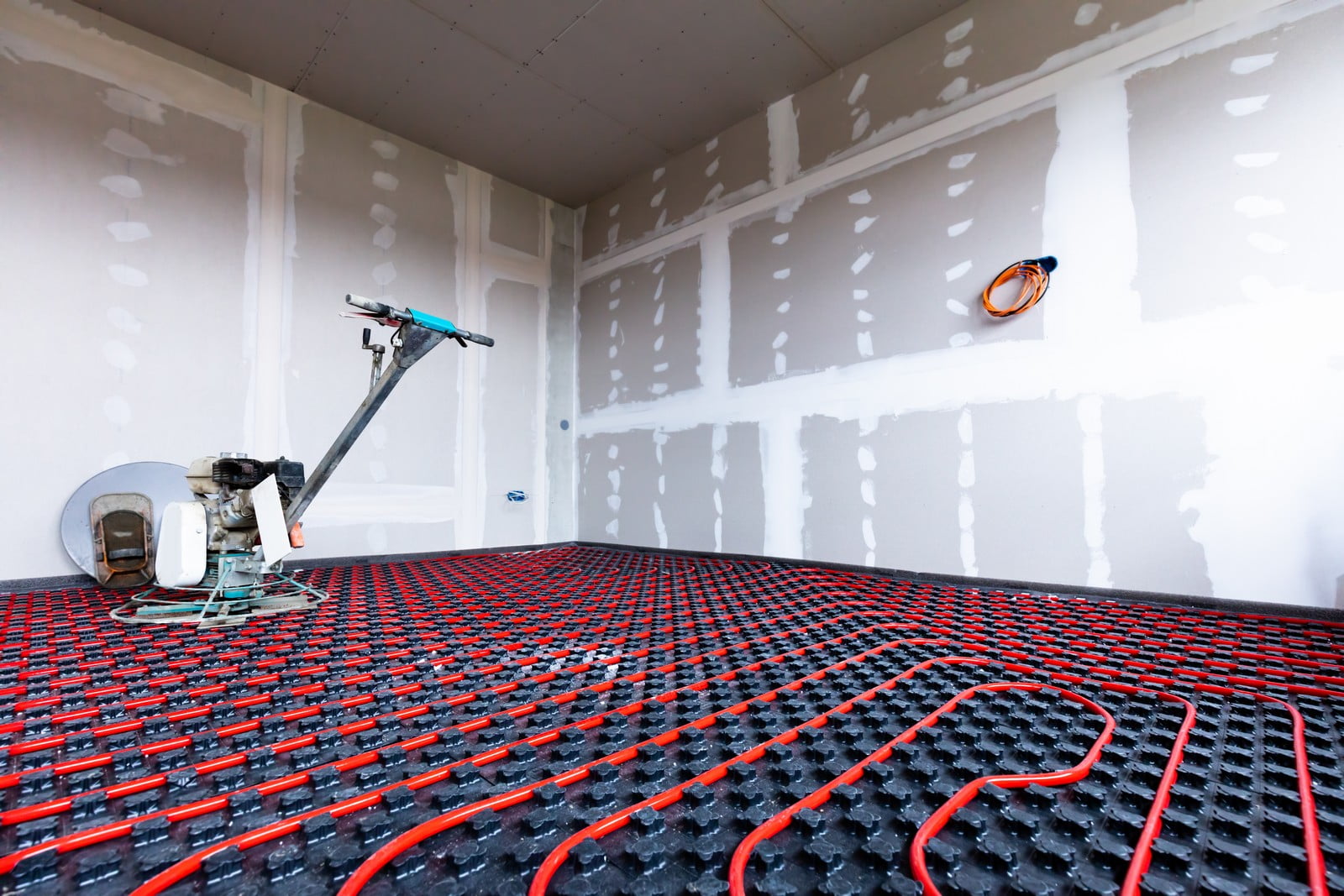 The image shows an under-construction or renovation space with an in-floor radiant heating system installed. There's red piping laid out across the floor in an evenly spaced pattern, which will circulate hot water or another fluid to heat the space from below. This layout is on top of what appears to be a dark grid-like mat that may provide insulation or support for the pipes.The walls are unfinished, with drywall installed but not yet painted—the seams and screw holes are covered with joint compound that hasn't been sanded down, which is a step in preparing walls for painting.On the left, there's some kind of machine or tool with a handle that resembles a scooter or manual tile cutter, but without additional context, it's not clear what its exact purpose is. There are no visible persons or identifiable personal items in the image, which suggests that work is not actively being done at the moment the photo was taken.