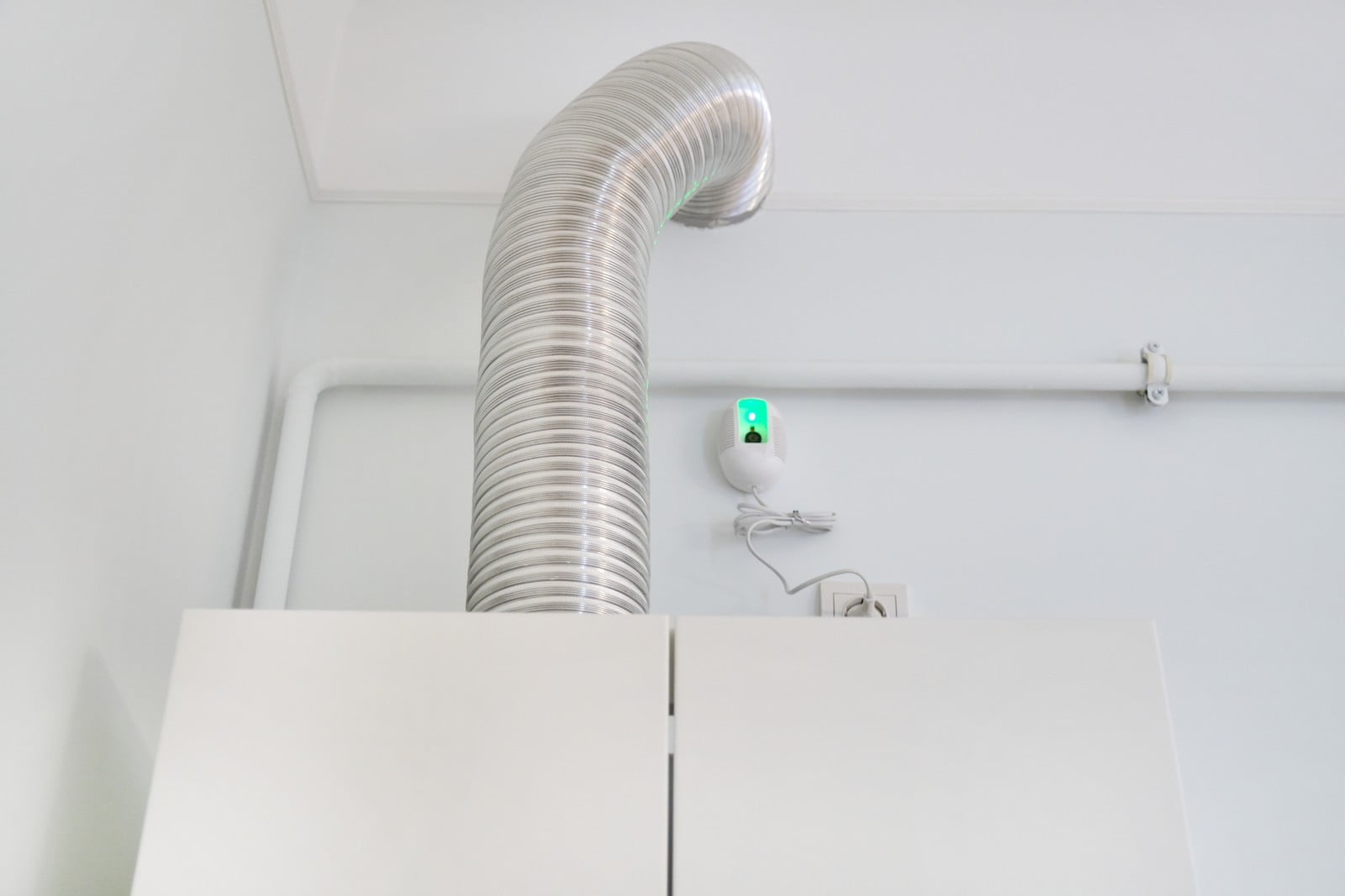 The image appears to show a section of an interior space with an HVAC (heating, ventilation, and air conditioning) duct or flexible aluminium hose extending from the ceiling. It seems to be attached to some sort of system for air circulation, extraction, or temperature control.One the right side of the image, there is an electrical outlet with a device plugged in that has a green illuminated indicator, suggesting that the device is powered on. The exact function of the plugged-in device isn't clear from the image, but it could be a sort of air freshener, night light, or another electrical appliance.Also visible are white cabinets or storage units at the bottom, and what looks like a white pipeline or conduit running horizontally along the wall, likely for electrical wiring or plumbing. The environment suggests a functional, utilitarian space such as a kitchen, laundry room, or industrial area. The overall setting seems clean and well-maintained, with a minimalist and modern aesthetic.