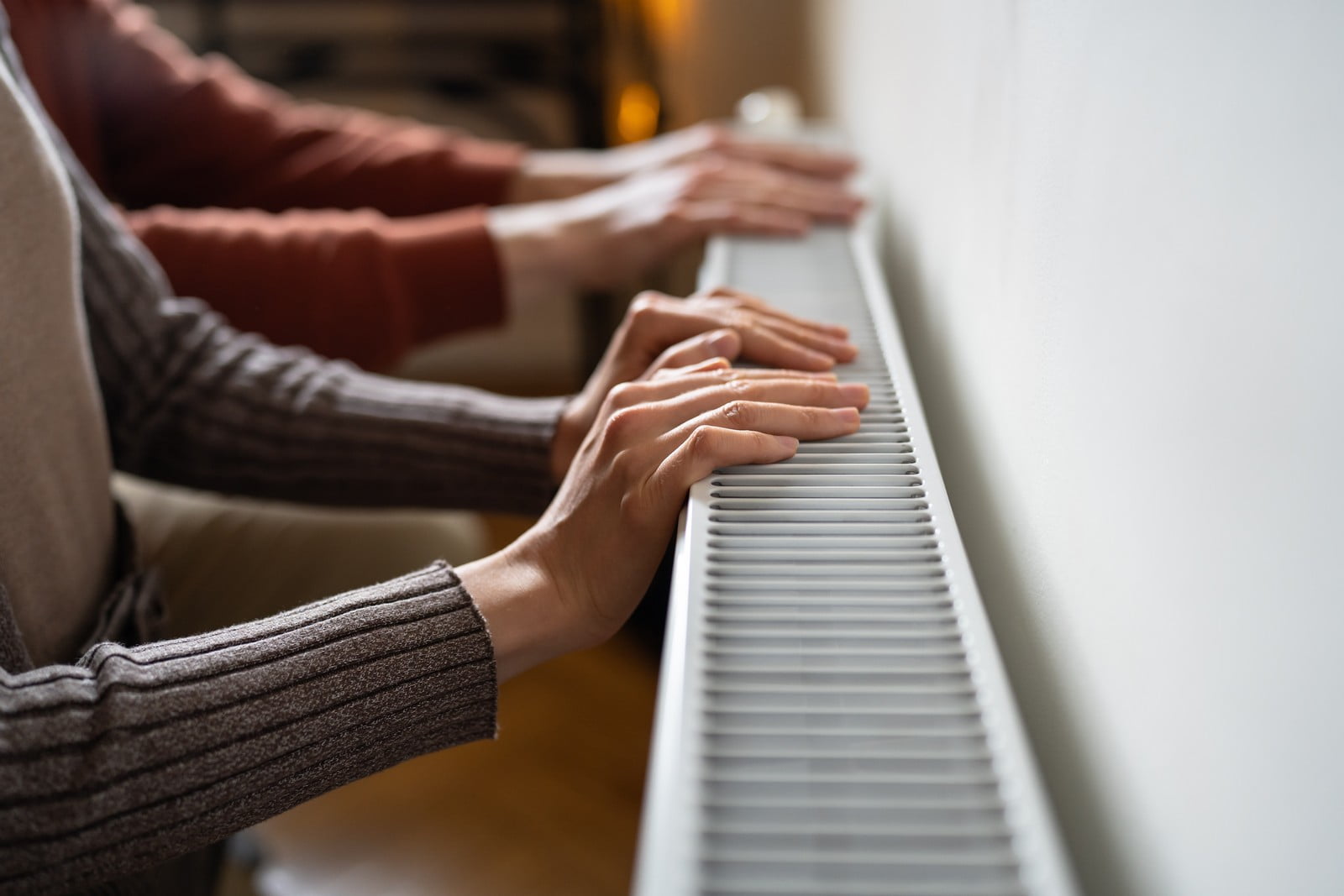 The image shows multiple people with their hands placed on what appears to be a radiator. They might be warming their hands. The radiator is mounted on a wall, and part of the room is visible, suggesting an indoor setting. It looks like a cold day, which is why the individuals are seeking warmth from the heating system. The focus of the photo is on the hands and the radiator, indicating the primary action or theme of the image.