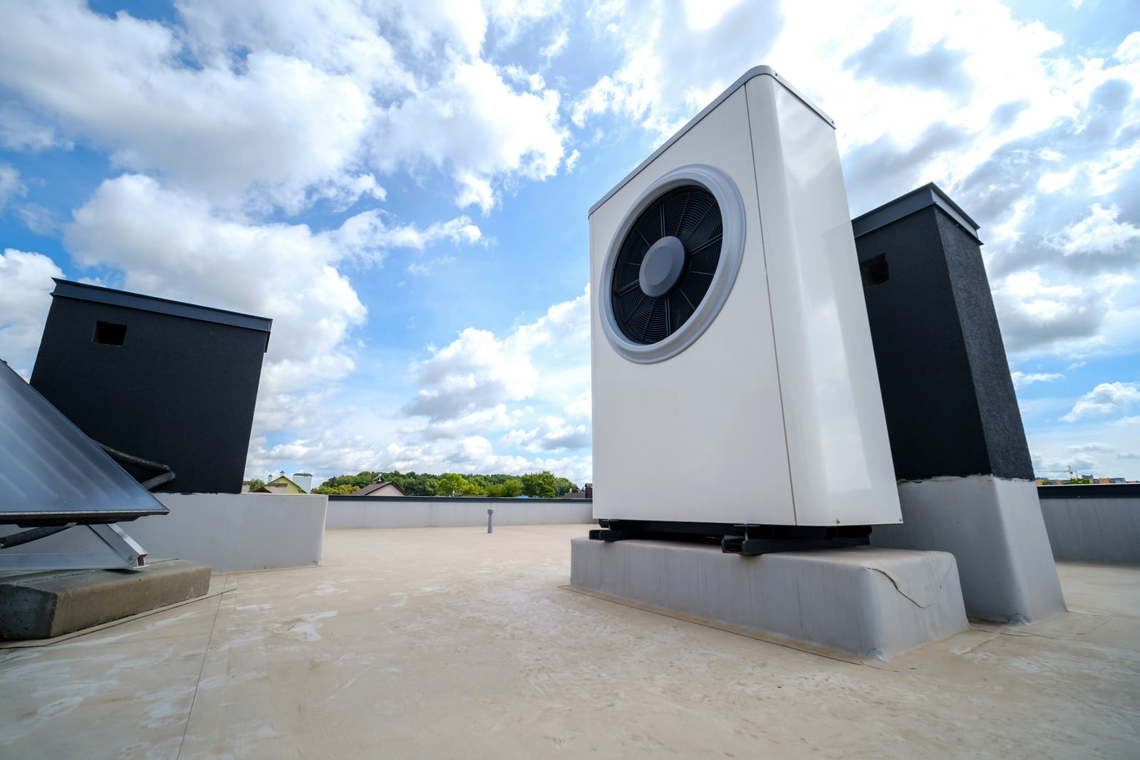 The image shows what appears to be a rooftop with various installations. Prominently, there is a large air conditioning or HVAC unit with a big fan visible; it is the central focus of the photo. The unit is placed on a platform raised above the roof surface, likely for maintenance access and to protect the roof membrane. Around this central unit, there are other less prominent features such as a skylight, some sort of vent pipes, and a couple of dark-coloured structures that might be access points, additional HVAC equipment, or even parts of a rooftop mechanical room. The sky is partly cloudy, suggesting a day with intermittent sunshine.