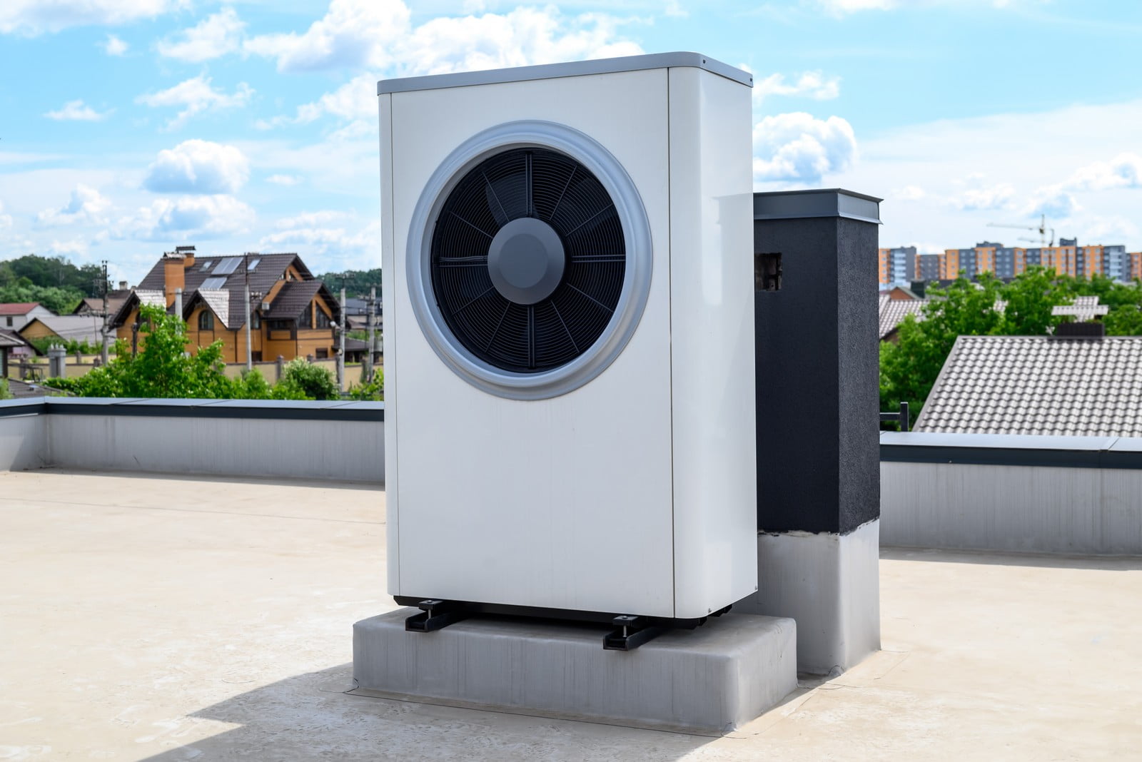 This image shows an outdoor unit of an HVAC system (Heating, Ventilation, and Air Conditioning) installed on top of a flat roof. The unit is likely to be an air conditioner or heat pump condenser part of a split system, where such units are installed outside and work in conjunction with an indoor unit. The background provides a view of a residential area with houses, greenery, and farther away, some flat buildings under a partly cloudy sky.