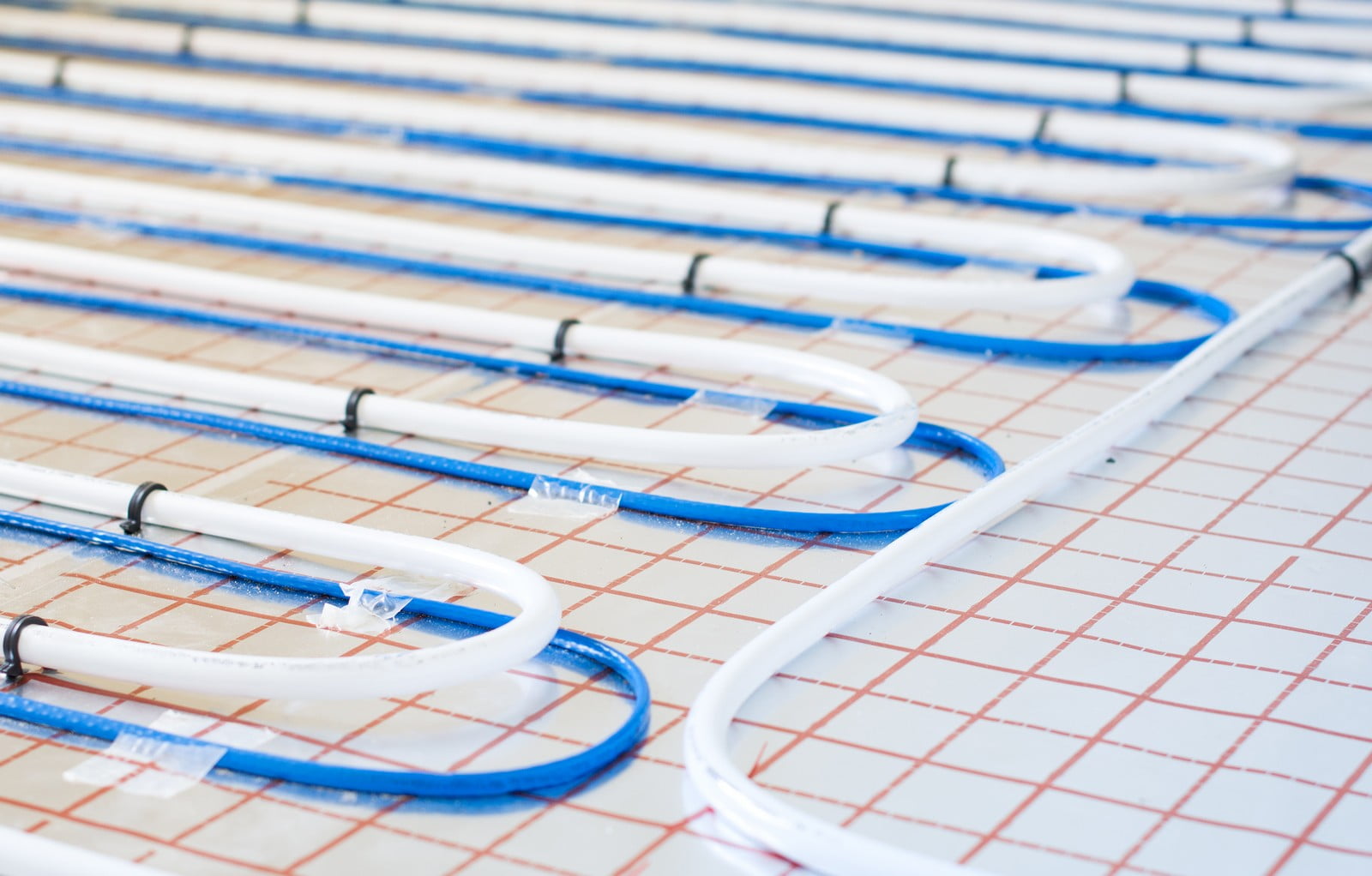 This image shows a close-up of a radiant floor heating system before the final flooring is laid over it. The white and blue pipes are arranged in a serpentine pattern to distribute hot water evenly beneath the floor surface, which provides heating for the room. The pipes are secured to the floor, and you can see the grid on the underlay, which might be used as a guide for laying the pipes with consistent spacing for optimal heat distribution.