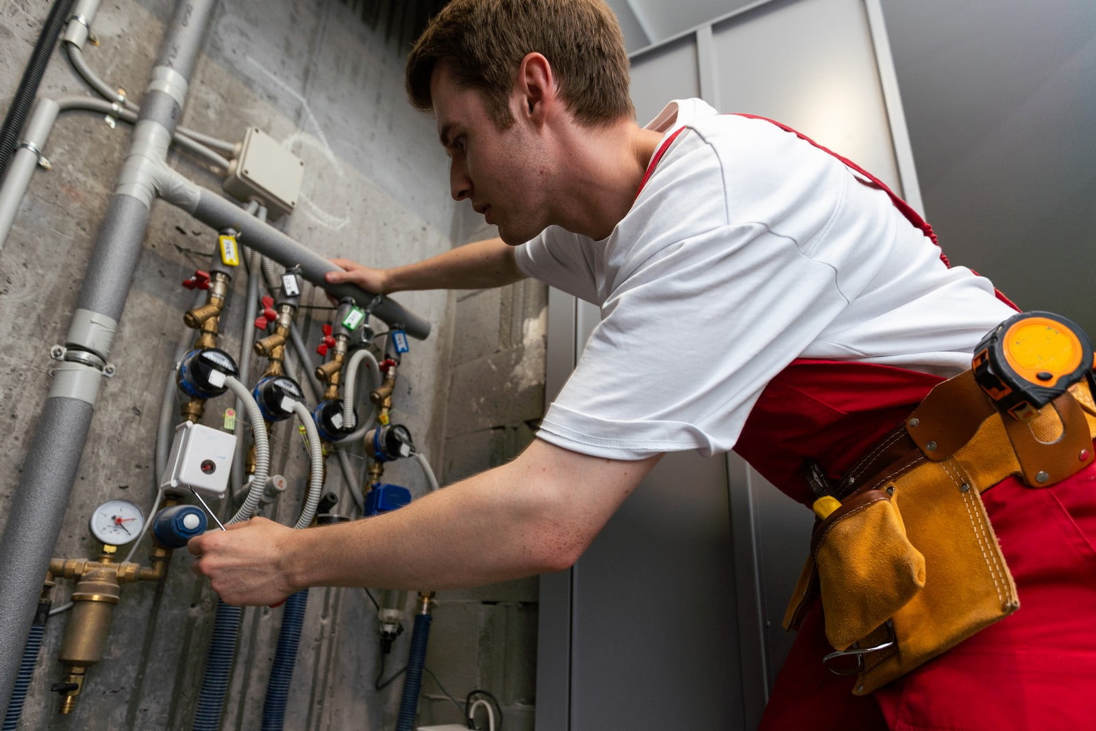 The image shows a man working on an industrial piping system or HVAC equipment. He is wearing a white t-shirt and red work pants with a yellow-and-brown tool belt around his waist. The tool belt holds various items, possibly tools or equipment necessary for his job. He appears to be adjusting or inspecting valves or gauges. There are multiple pipes, valves, sensors, and a pressure gauge in the setup, which could be part of a heating, ventilation, and air conditioning system. The environment suggests it could be in a maintenance room or a utility area of a building.