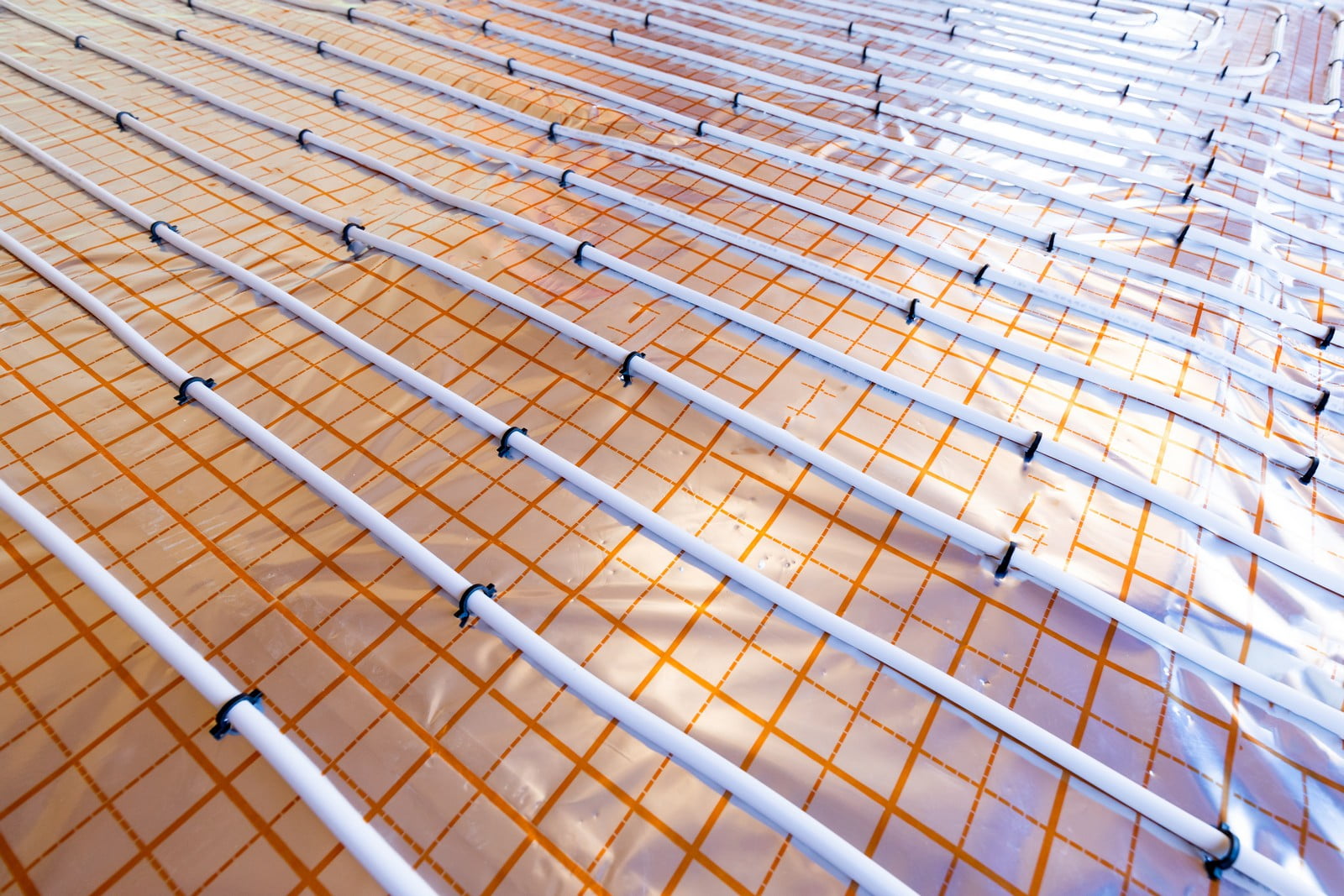 The image shows an underfloor heating installation before the final flooring is applied. You can see a series of pipes laid out in parallel runs across the floor. These pipes will circulate warm water, which heats the floor and then radiates heat into the room above. The orange grid beneath the pipes is likely insulation intended to direct the heat upward and prevent heat loss to the ground. Underfloor heating is an efficient method for creating a comfortable indoor climate, especially in cooler climates where radiant floor heating can provide an even, gentle warmth.