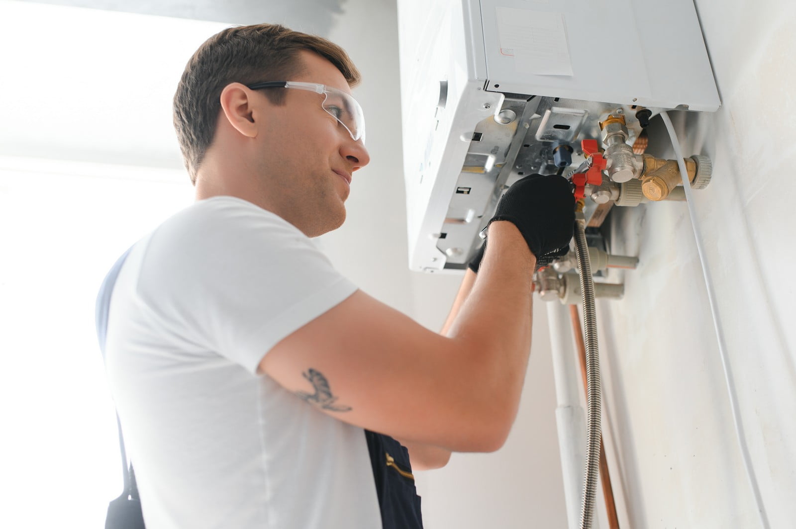 This image features a person working on a wall-mounted petrol boiler or water heater. The person is likely a technician or a plumber as suggested by the workwear, including a white t-shirt, safety goggles, gloves, and a tool belt or work pants. The individual is focused on the task and appears to be adjusting or inspecting the internal components of the boiler, which are visible with the cover panel removed. There are various pipes and valves connected to the appliance, indicating it's part of a larger heating or hot water system for a building.