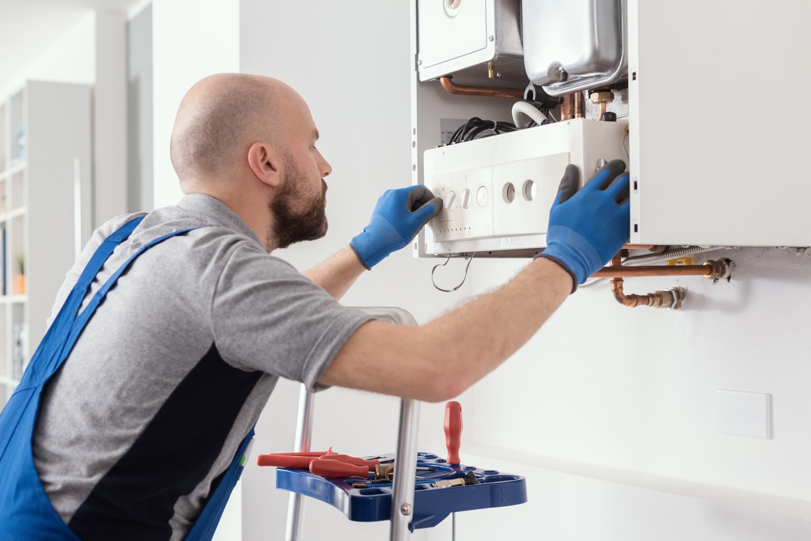 The image shows a person performing maintenance or repair work on a domestic boiler or heating system. The individual is wearing a gray shirt, blue overalls, and blue gloves. He is working attentively on the internals of the boiler system, which is mounted on a white wall. The front panel of the boiler is open, allowing access to its components. Below the boiler, copper pipes can be seen connecting to the central heating system.The workman has a tool bag or box with various tools like pliers and screwdrivers on a small stepladder beside him, which suggests preparedness for various tasks related to the repair or maintenance job at hand. It appears to be an indoor setting, presumably within a home or residential building. The worker's attention to the system indicates professionalism and focus on ensuring the proper functioning of the boiler.