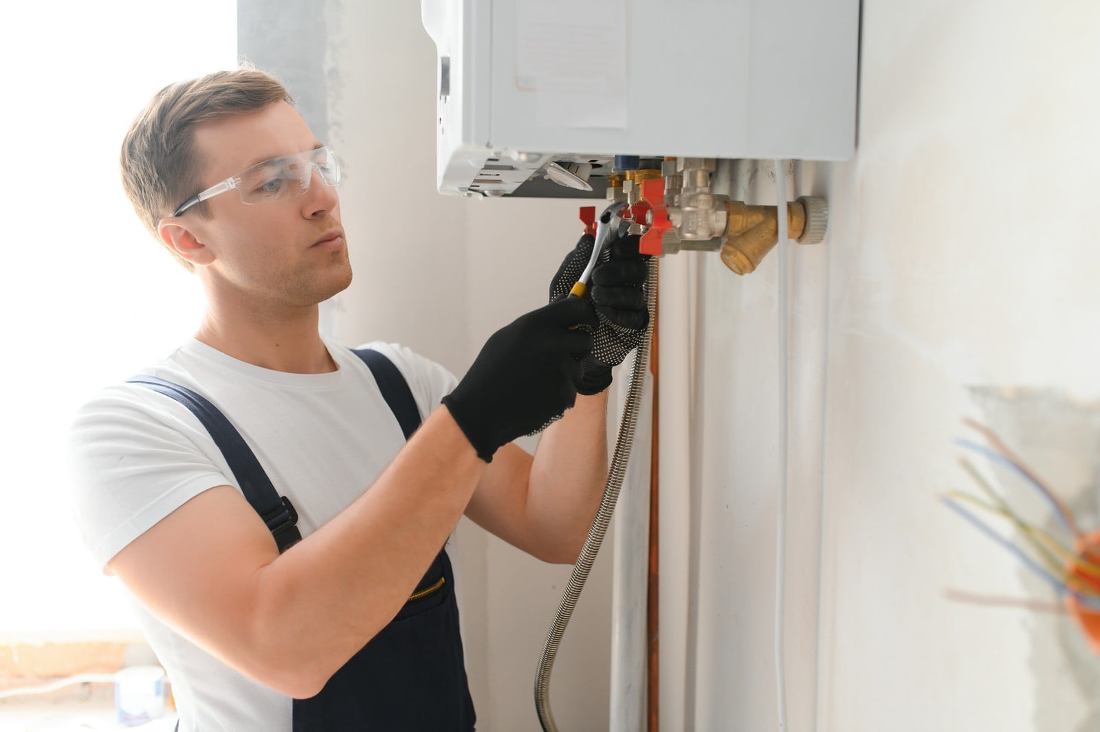 This is an image of a person who appears to be a technician or plumber installing or repairing a water heater or boiler. He is wearing protective eyewear and gloves as safety precautions. The technician is focused on his work, using a spanner to adjust or secure connections on the heater. The equipment he is working on is wall-mounted, and there are pipes and valves that are part of the installation. It's a clean and bright environment, suggesting that this might be a residential or commercial setting.