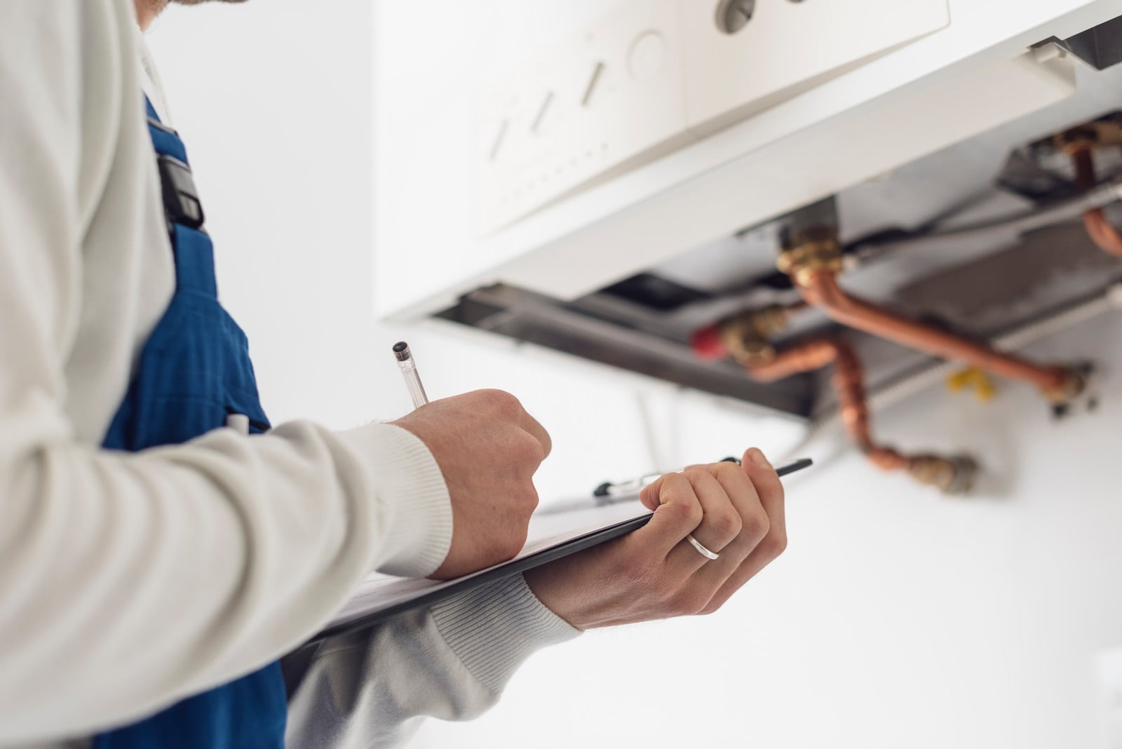 The image shows a person in a white long-sleeve shirt and wearing a blue work apron, likely a technician, holding a clipboard with a pen. They seem to be inspecting or servicing a wall-mounted heating appliance, which could be a boiler. Only part of the person's body is visible; the image focuses on the person's hands, the clipboard, and the appliance, which has copper pipes and valves visible, indicating it is part of a heating, ventilation, and air conditioning (HVAC) system.