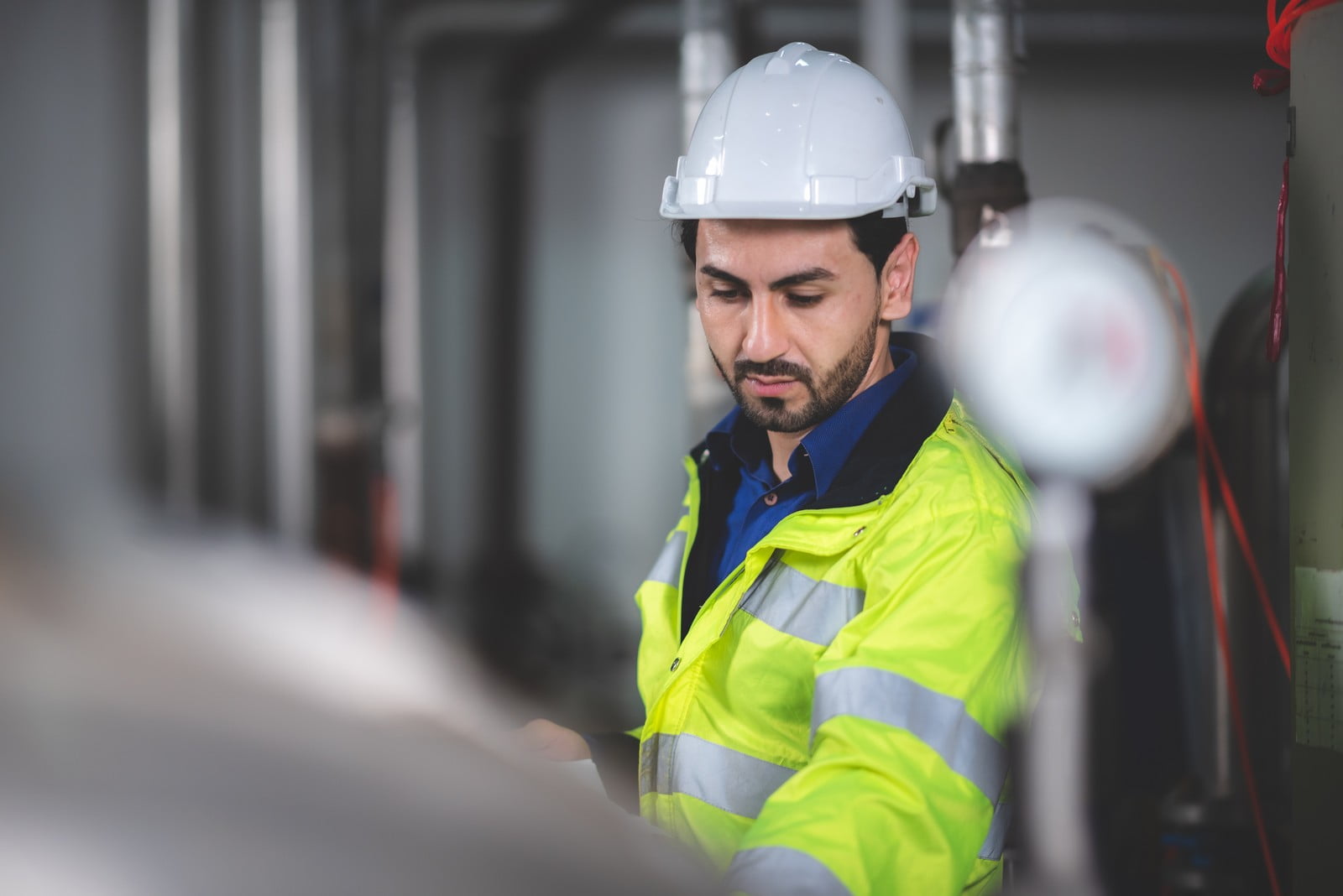 The image displays a person who appears to be a worker or engineer. The individual is wearing a safety helmet and a high-visibility jacket with reflective stripes, which are common safety garments in industrial or construction environments. The worker is focused on something off-camera, and there seems to be industrial equipment in the blurred background, suggesting the setting might be a factory, plant, or other industrial site.