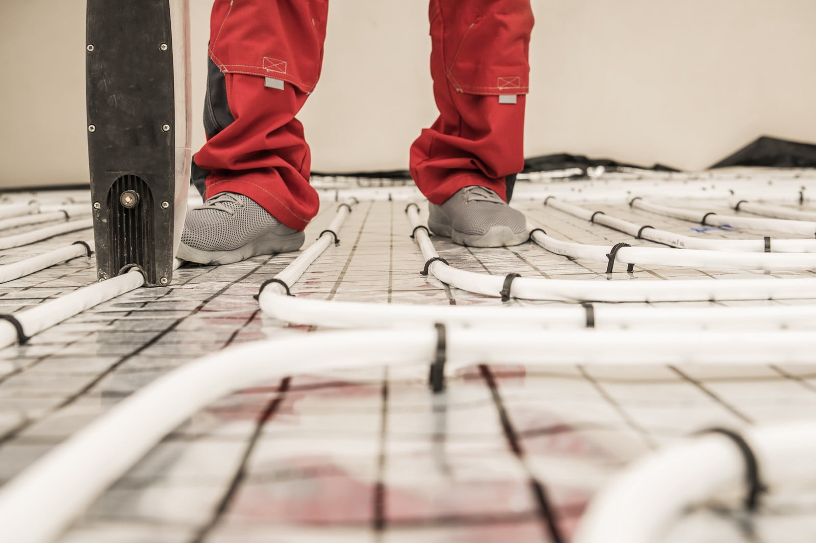 This image shows a close-up of a person standing amidst what appears to be underfloor heating installation. The heating system consists of pipes laid out across the floor, which will likely be covered with a finished flooring surface after installation. The person is wearing red work trousers, and the black parts on the trousers near the knees suggest they might be reinforced for kneeling, which is common in workwear for construction or installation jobs. There's also a ladder seen in the photo, which the person might use for their work. The floor is still exposed, indicating that the installation process is not yet complete.