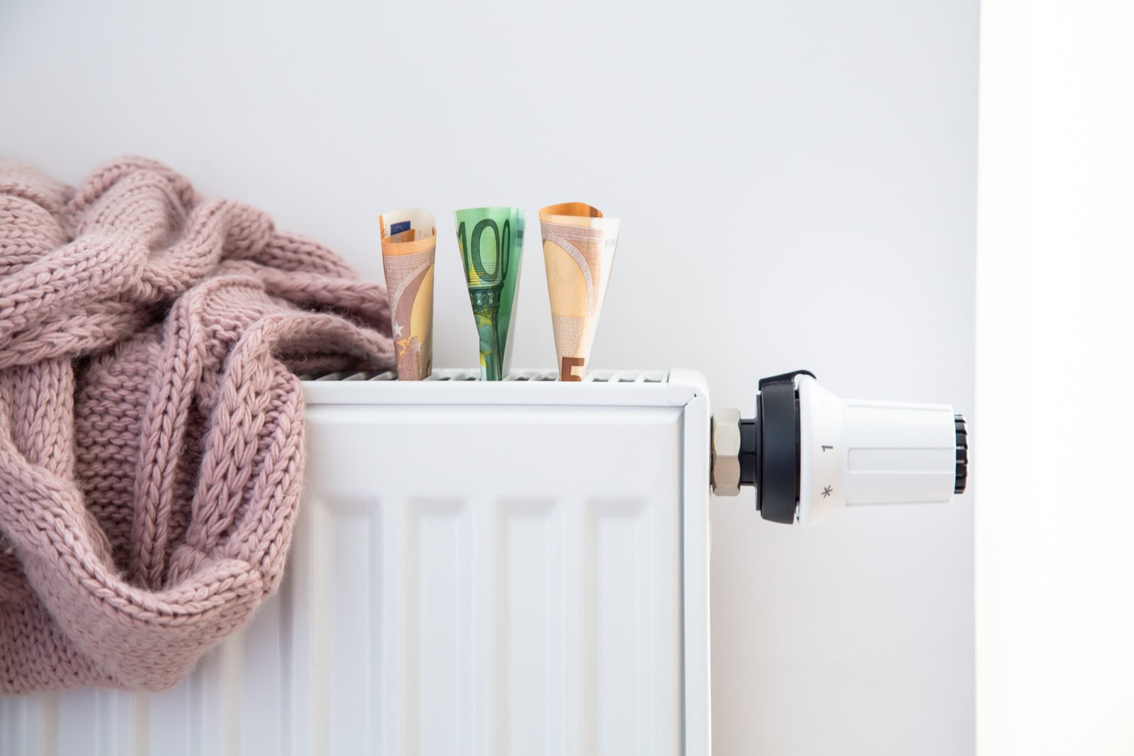 This image shows a white radiator with a pink knitted blanket draped over it. On top of the radiator, there are three rolled-up Euro banknotes -- a fifty, a ten, and a five. To the right of the radiator is a thermostatic radiator valve, which is used to control the temperature of the radiator. The setting of the room appears to be a domestic interior, likely aimed at demonstrating a concept related to saving money on heating or the cost of heating a home.