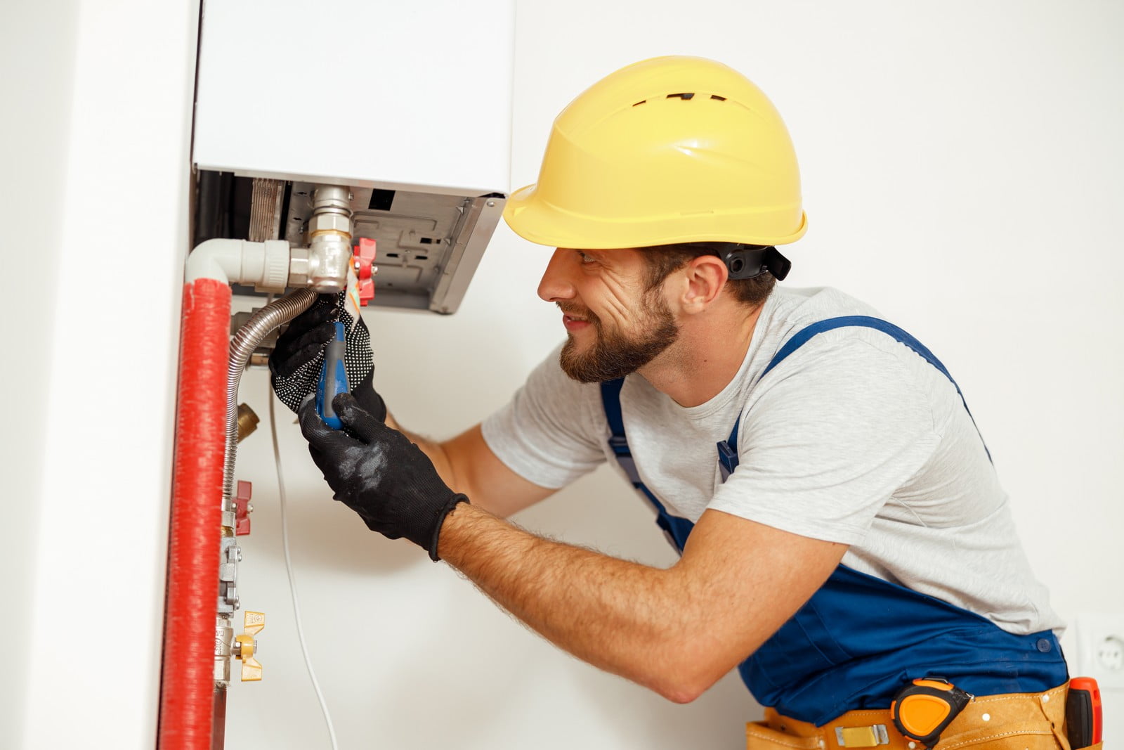 The image shows a man working on a piece of equipment that appears to be a domestic boiler or a heating system component. He is wearing a yellow hard hat, suggesting adherence to safety standards, and has protective gloves on. His tool belt suggests he is a professional technician or maintenance worker. He's holding a flexible hose and examining the interior part of the equipment, possibly performing maintenance, repair, or installation work. The environment looks clean and residential.