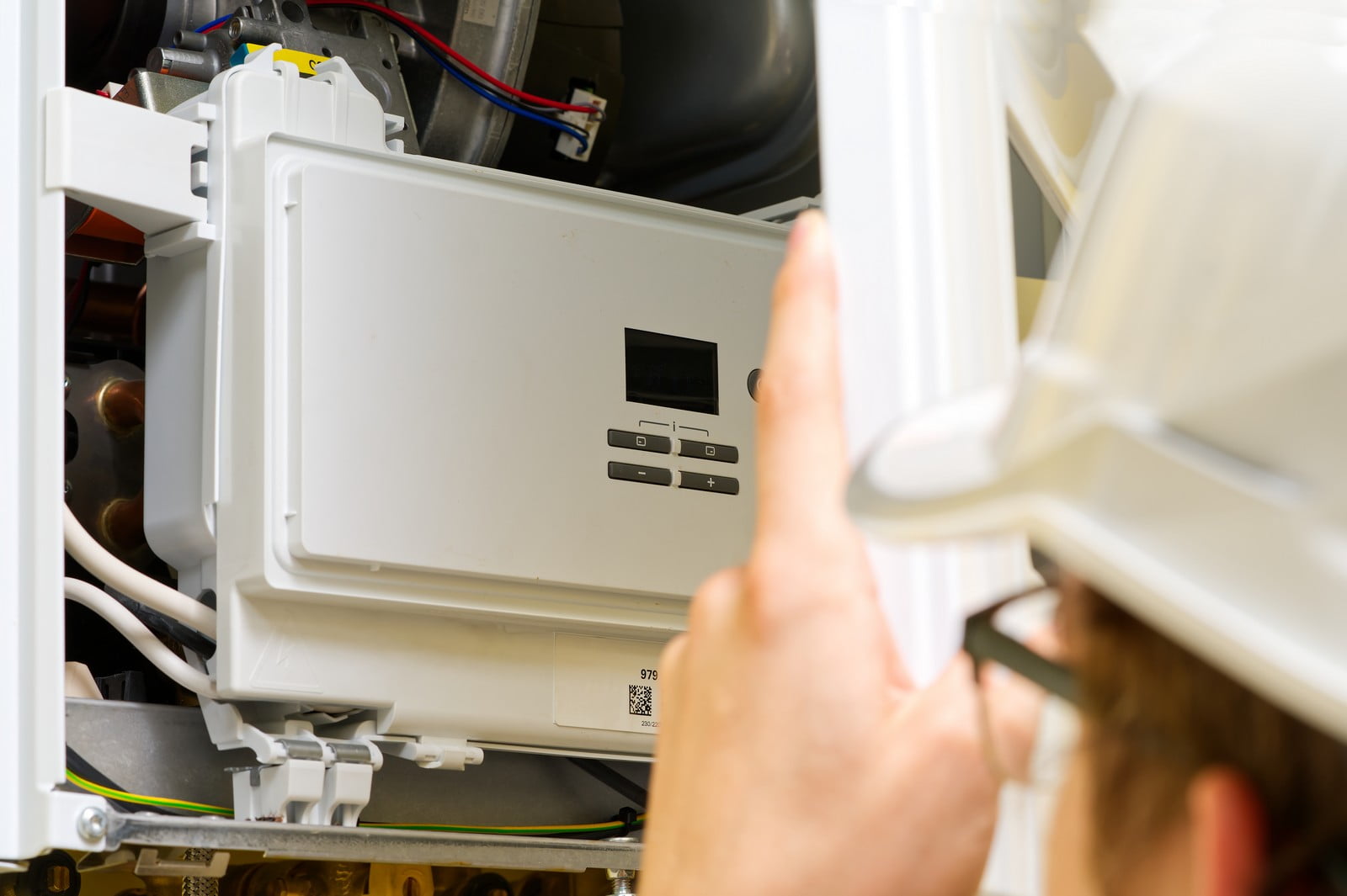 This image shows a person, likely a technician or engineer, who is working on what appears to be a domestic appliance, possibly a boiler or a heating, ventilation, and air conditioning (HVAC) unit. The person is wearing a hard hat, indicating they are adhering to safety standards typically required in maintenance or construction settings.The interior of the appliance is partially visible, with electronic components and wiring suggesting that this is a control panel the technician is interacting with. There are buttons and a digital display on the white panel, which might be used to set or monitor the appliance's operation or to troubleshoot issues.The focus of the image is on the control panel and the person's hand as they reach into or interact with the unit, with the rest of the appliance and the person's body being out of focus. This kind of maintenance or inspection is essential for the safe and efficient operation of such household appliances.