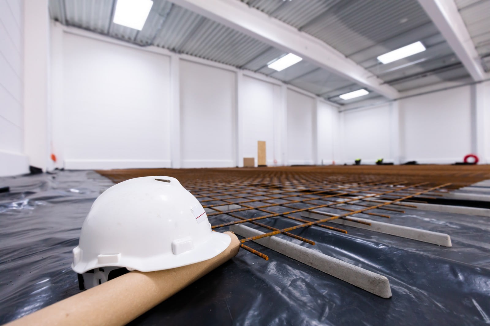 The image shows an indoor construction site. In the foreground, there is a white hard hat lying on its side, a common piece of personal protective equipment used on construction sites to protect workers from head injuries. It's resting on what appears to be a roll of paper or building plans.Beneath the hard hat and covering the floor is a black plastic sheet, which is likely used as a vapour barrier or to keep the site clean. In the middle ground, there is a network of steel rebars laid out in a grid pattern, indicating that this is likely the preparation for a concrete pour for a foundation or floor slab.There are also a couple of trowels on the plastic, which are tools used for spreading, smoothing, leveling, or shaping concrete. In the background, the environment looks like a large, open space with white walls and high ceilings with skylights providing natural light. There are a few more elements of construction visible in the distance, but the details are indistinct. The overall impression is that of an active construction site where the foundation or flooring work is in progress.