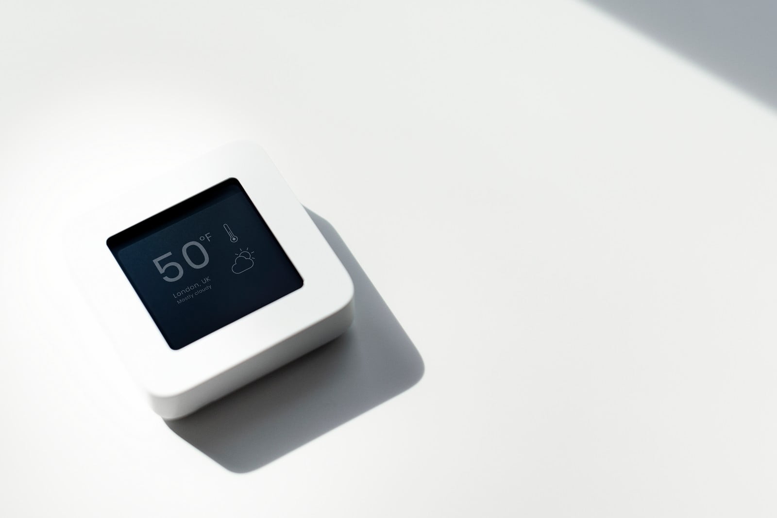 The image shows a digital thermostat on a light-coloured surface. The thermostat has a simplistic, modern design with a white casing, and its display is indicating a temperature of 50 degrees Fahrenheit. There is also a weather icon suggesting partly cloudy conditions, and the location is specified as London, UK. The background of the image is clean and minimalist, primarily white, which gives the impression of a bright, open space. The play of light and shadows adds a subtle depth to the composition.