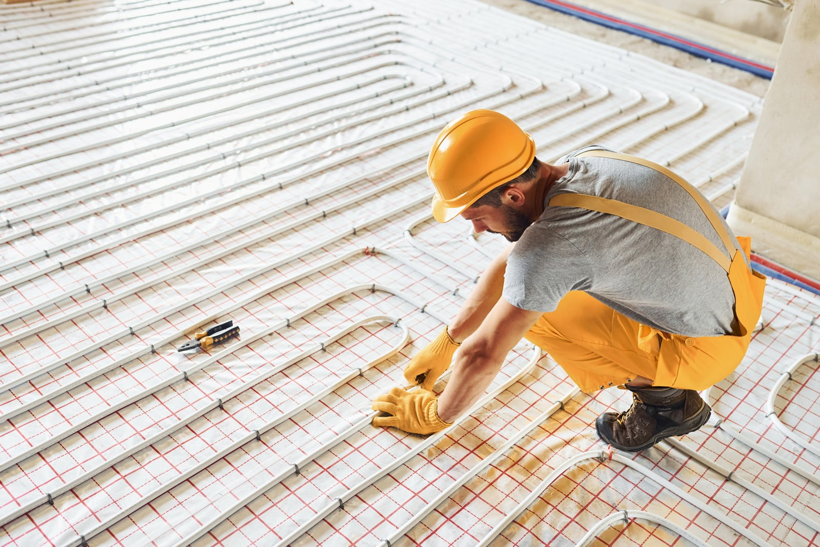 The image shows a person working on the installation of an underfloor heating system. The individual is wearing a yellow hard hat and gloves for safety, as well as yellow work pants with braces and sturdy work boots. They appear to be engaged in installing or inspecting the pipes that are a part of the heating system, which are laid out on the floor in a pattern to distribute heat evenly once completed. The pipes are secured to the insulating underlayment, which has grid lines to help with the placement of the pipes. There is also a tape measure beside the worker, suggesting precise measurements are being taken during the installation process. The setting suggests that this is a construction or renovation site.