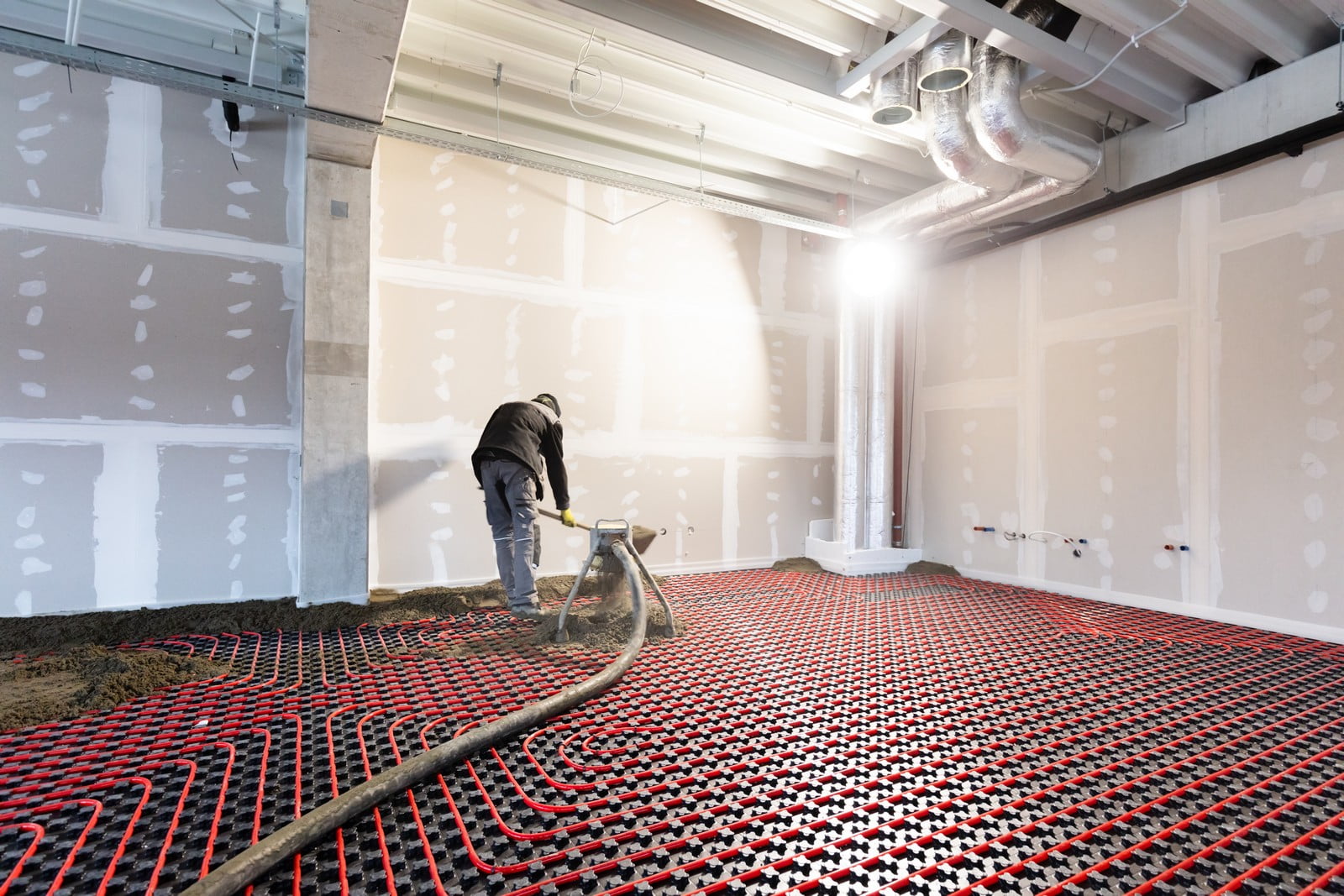 The image shows an interior construction scene where a worker is involved in the process of laying down a radiant floor heating system. You can see the red pipes laid out in an organized, serpentine pattern across the floor, which will circulate warm water to heat the room evenly from the ground up. This system is installed before the final flooring is laid over it.The room looks like it's in an early stage of renovation or construction, with exposed walls that have been prepped with joint compound (often referred to as "mud") for drywall finishing, and visible ductwork and structural elements on the ceiling. The construction worker is using a trowel or similar tool to apply a layer of cement or self-leveling concrete over the heating pipes, which will encase the system before the final flooring is installed. The scene is brightly lit, probably by natural light coming through the windows, and by work lights as well.