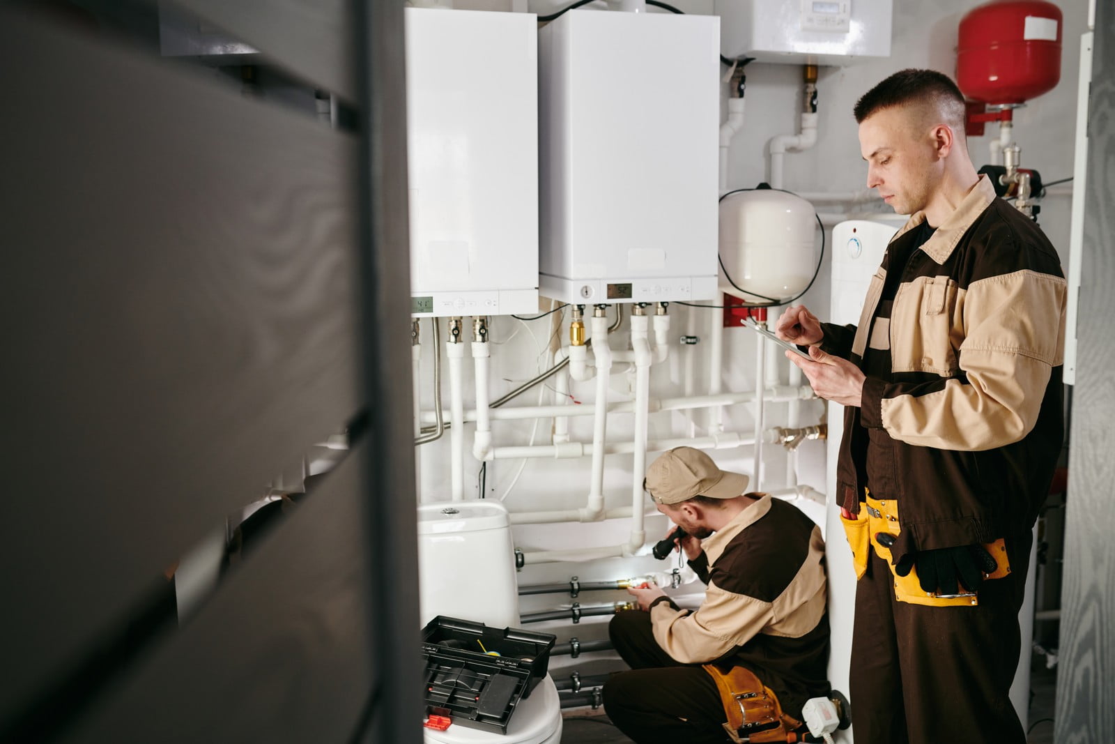 This image shows two technicians working on heating systems within a utility room. The one on the right is standing and using a mobile device, possibly reading or entering information related to the job. The other technician is crouched down, examining or working on part of the system with a torch. They are both dressed in work clothes with visible tool belts, and there are boilers, pipes, and a red expansion tank within the room, which indicates they might be servicing or installing heating, ventilation, and air conditioning (HVAC) components. There's also a toolbox opened on the floor, suggesting that they are actively engaged in maintenance or repair work.