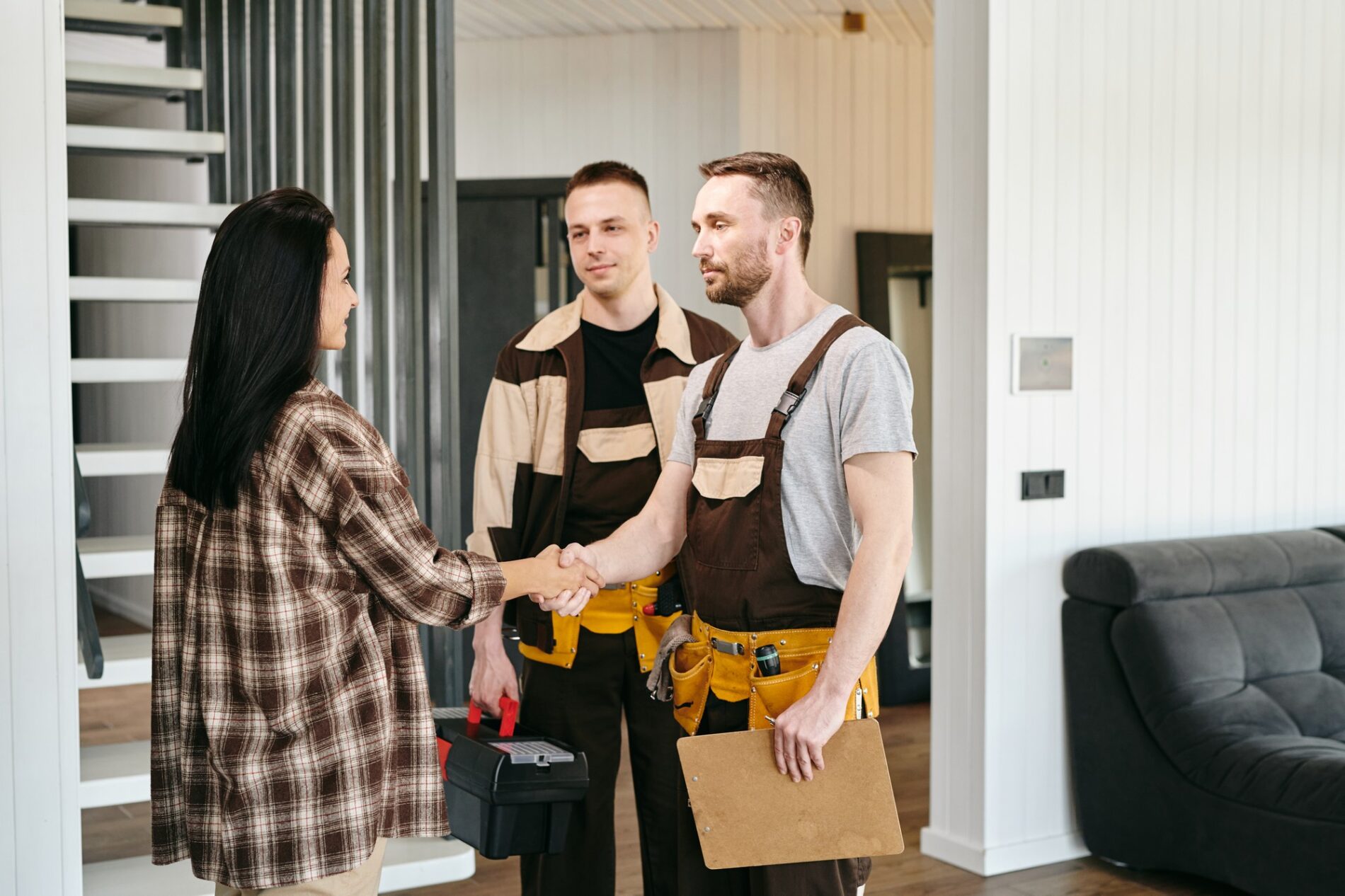 In the image, we see three individuals in what appears to be a home interior. A woman on the left is shaking hands with a man on the right, who is wearing a tool belt and carrying a clipboard, suggesting that he might be a tradesperson or contractor. Another man in the middle, standing slightly behind the one shaking hands, is also wearing a similar outfit and holding what looks like a toolbox, indicating he may also be a tradesperson or an assistant. The setting seems to be a professional visit, possibly for home repair or consultation. The home has a contemporary design with a staircase in the background as well as a comfortable-looking dark couch. Overall, the scene appears to depict a friendly or business interaction between a homeowner and one or more tradespeople.