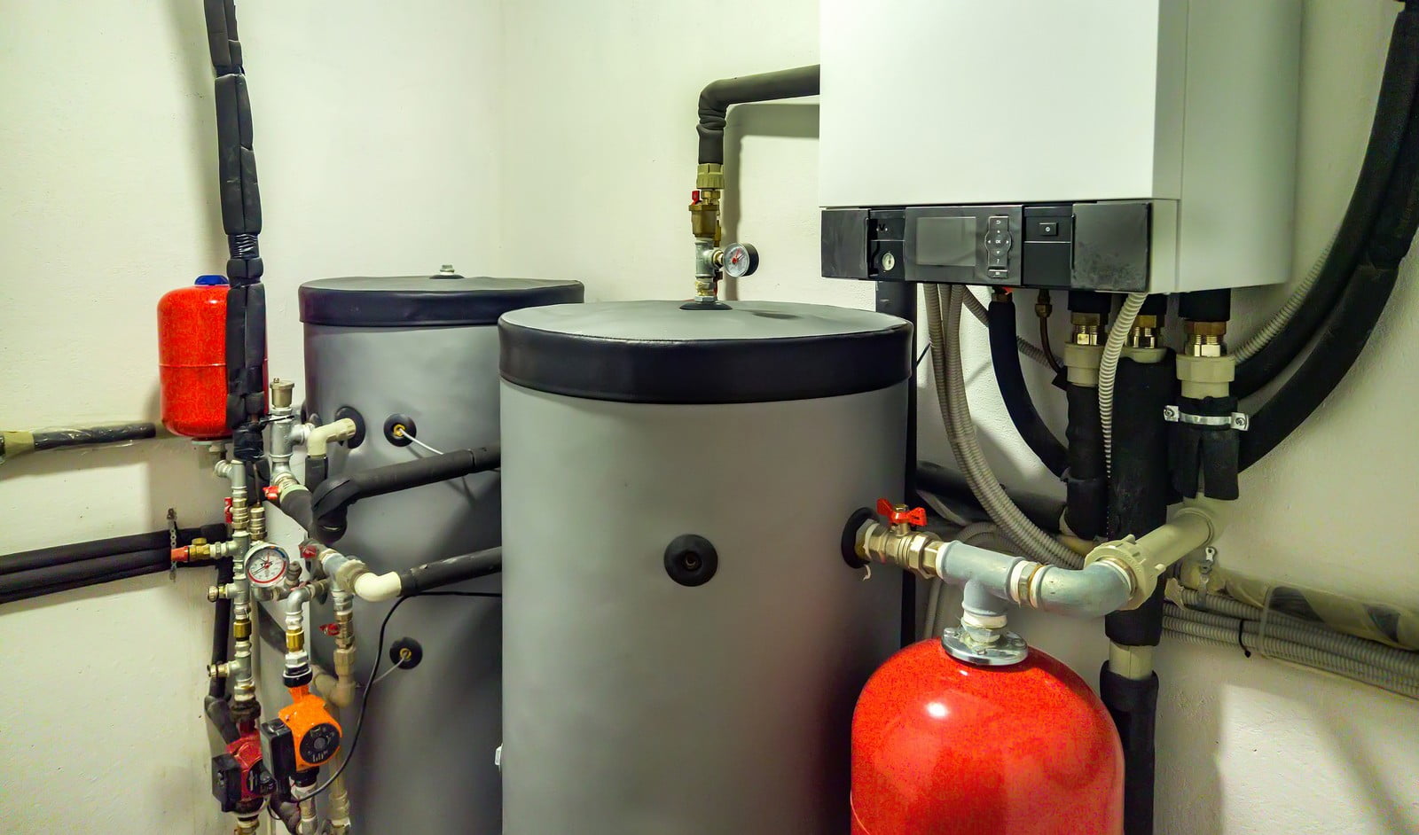 This image shows a domestic boiler room with various pieces of equipment that are part of a home heating system. On the right side, there's a wall-mounted petrol boiler, recognizable by its compact, boxy shape and control panel. The boiler has multiple pipes connected to it, suggesting it's integrated with the home's water supply for heating.There are two large tanks, likely expansion vessels, which are used to accommodate the expansion of water as it heats up in the system. These tanks have safety valves on top, which helps maintain pressure and prevent over-pressurization.Also visible are several pipes with insulation, valves, safety devices like pressure gauges, and possibly a red pressure relief valve connected to one of the tanks. The assortment of valves and connections indicates this is a well-established system, designed for efficiency and safety. The system also has electrical connections, and flexible hoses, indicating a modern setup.Maintenance components, such as the red expansion tank and various manual shutoff valves, are part of the system as well, suggesting this setup is organized for both operation and maintenance ease. The overall condition appears clean and well-maintained.