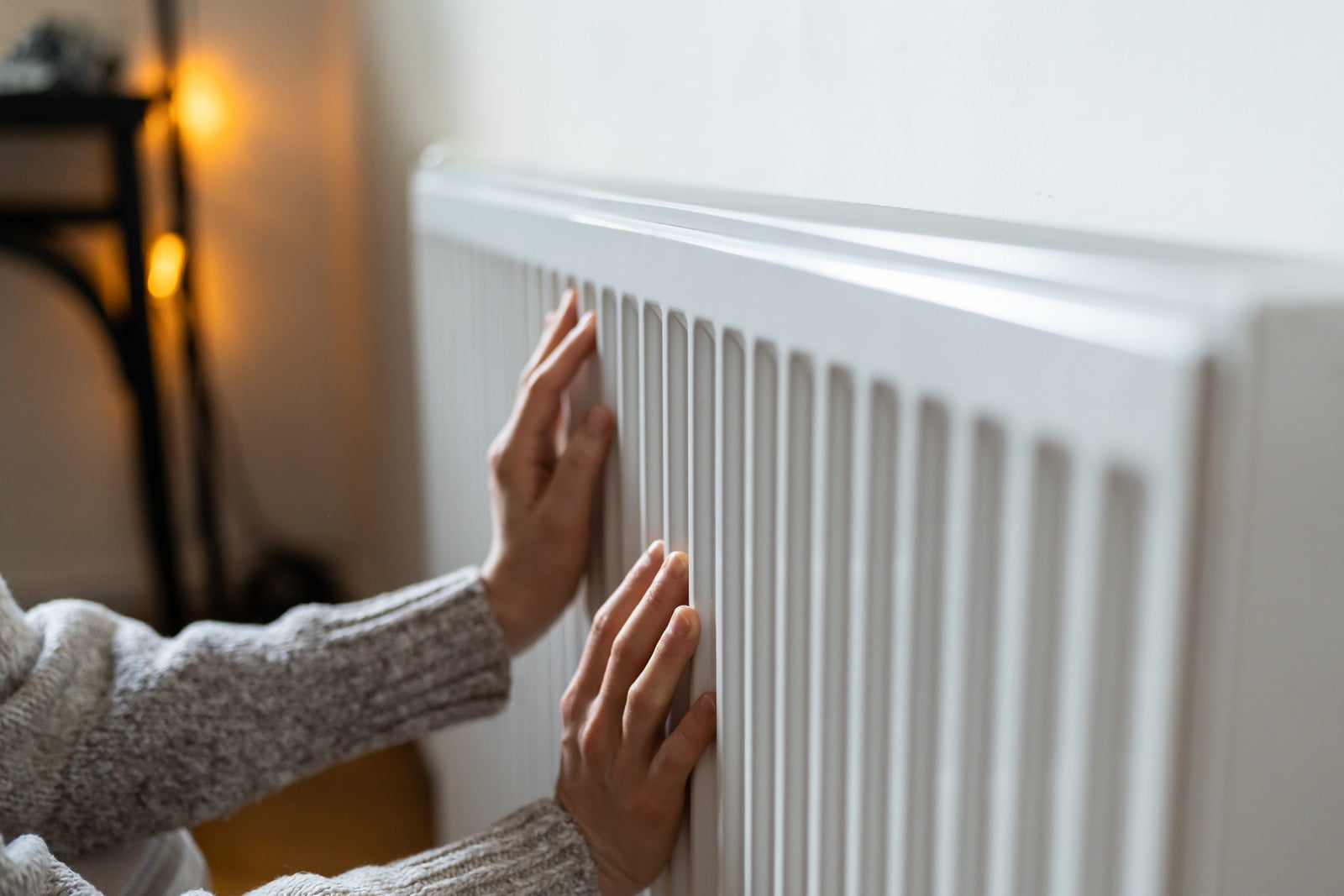 The image shows a person touching a white radiator, likely checking if it is warm. The individual is wearing a grey jumper, and only the lower part of their face and hands are visible, with a focus on the hand touching the radiator. The background is blurred, but it appears to be an indoor setting with a calm, warm ambiance suggested by soft lighting, possibly from a lamp, in the top left corner. The wall behind the radiator is white, providing a clean and simplistic background for the subject of the photo.