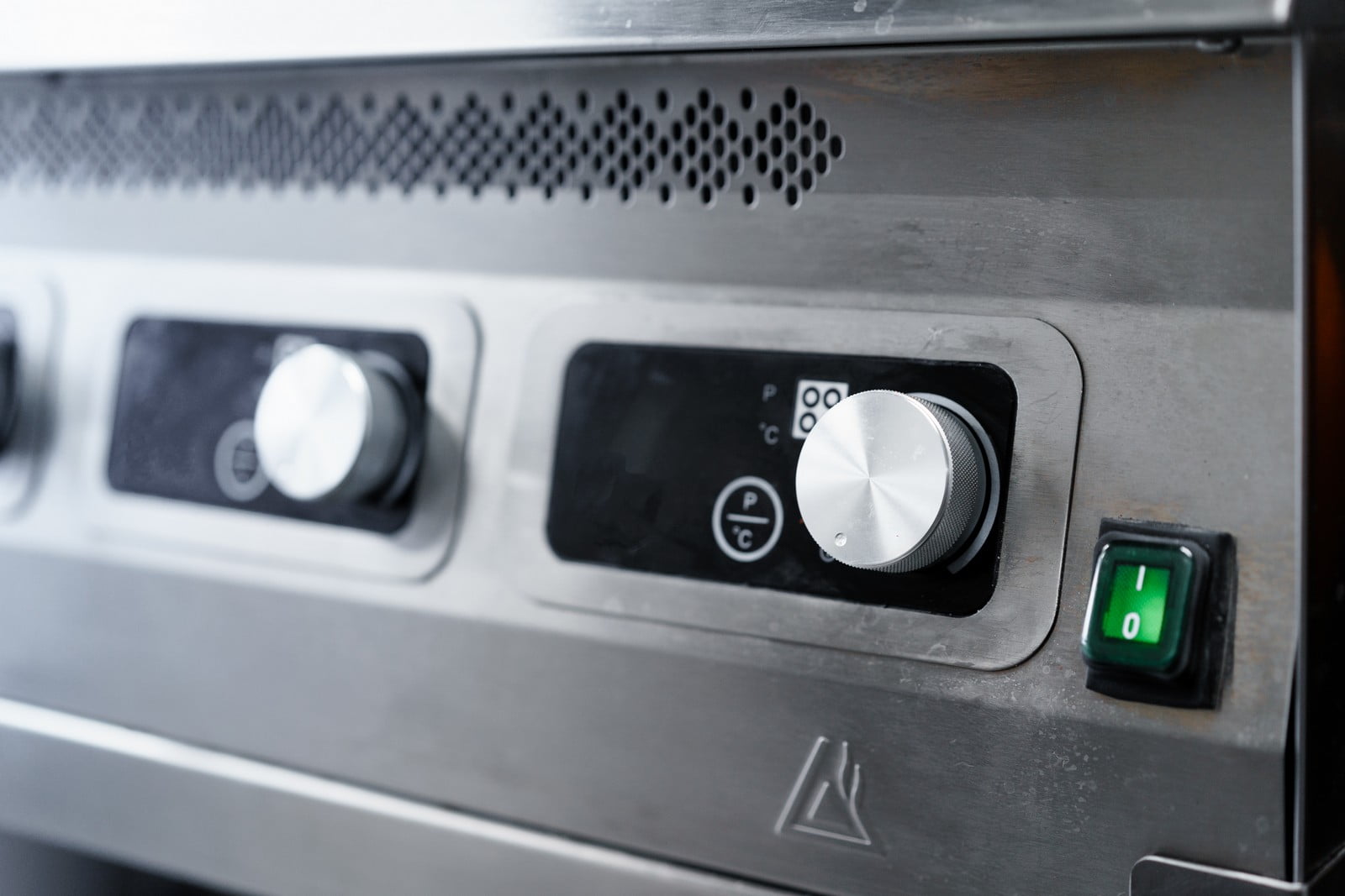 The image shows a close-up of a control panel, likely on a commercial kitchen appliance, such as an oven, range, or other cooking device. The panel includes a knob for adjusting settings, which appears to be made of metal with a textured edge to aid grip, and a digital display screen which might show temperature, time, or other operational information. To the right of the knob, there is a green-lit power switch indicating that the appliance is turned on. The stainless steel construction suggests it's designed for durability and ease of cleaning, common in professional kitchen environments.