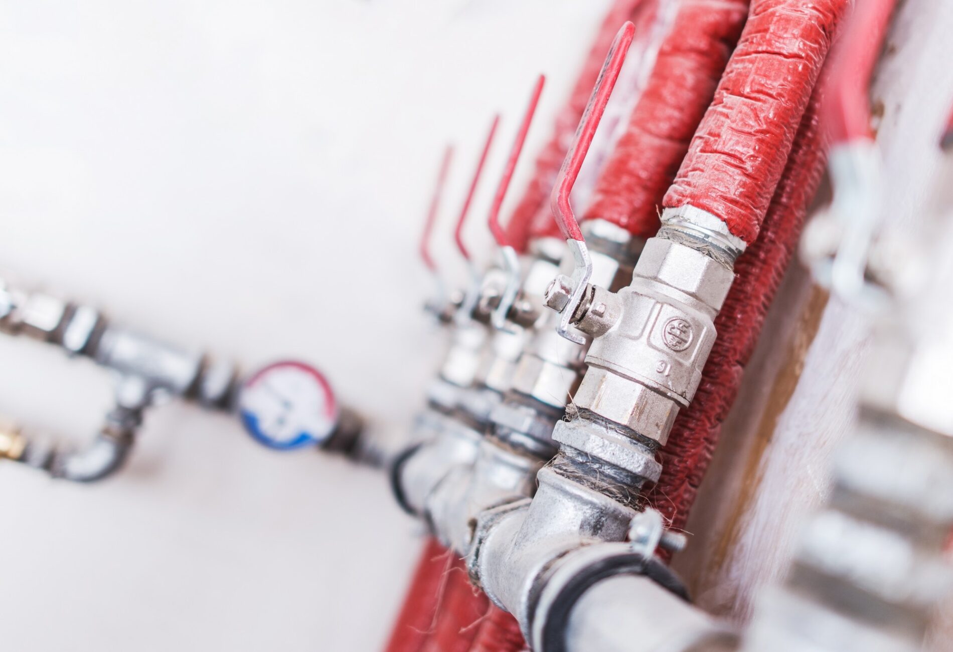 The image shows a series of metal pipes and valves, partially wrapped in what appears to be red insulation material. On each valve, there is a red handle that is used to open and close the valve, controlling the flow of whatever medium runs through the pipes. There's also a pressure gauge visible at the bottom left of the picture with a red and blue area on its dial, likely indicating pressure levels or temperatures. The focus of the image is shallow, with only a portion of the nearest valve and insulation in clear detail, and it gradually blurs out towards the background, giving a sense of depth. This setup is typical for industrial or commercial settings where temperature control or regulation of fluids or gases is necessary.