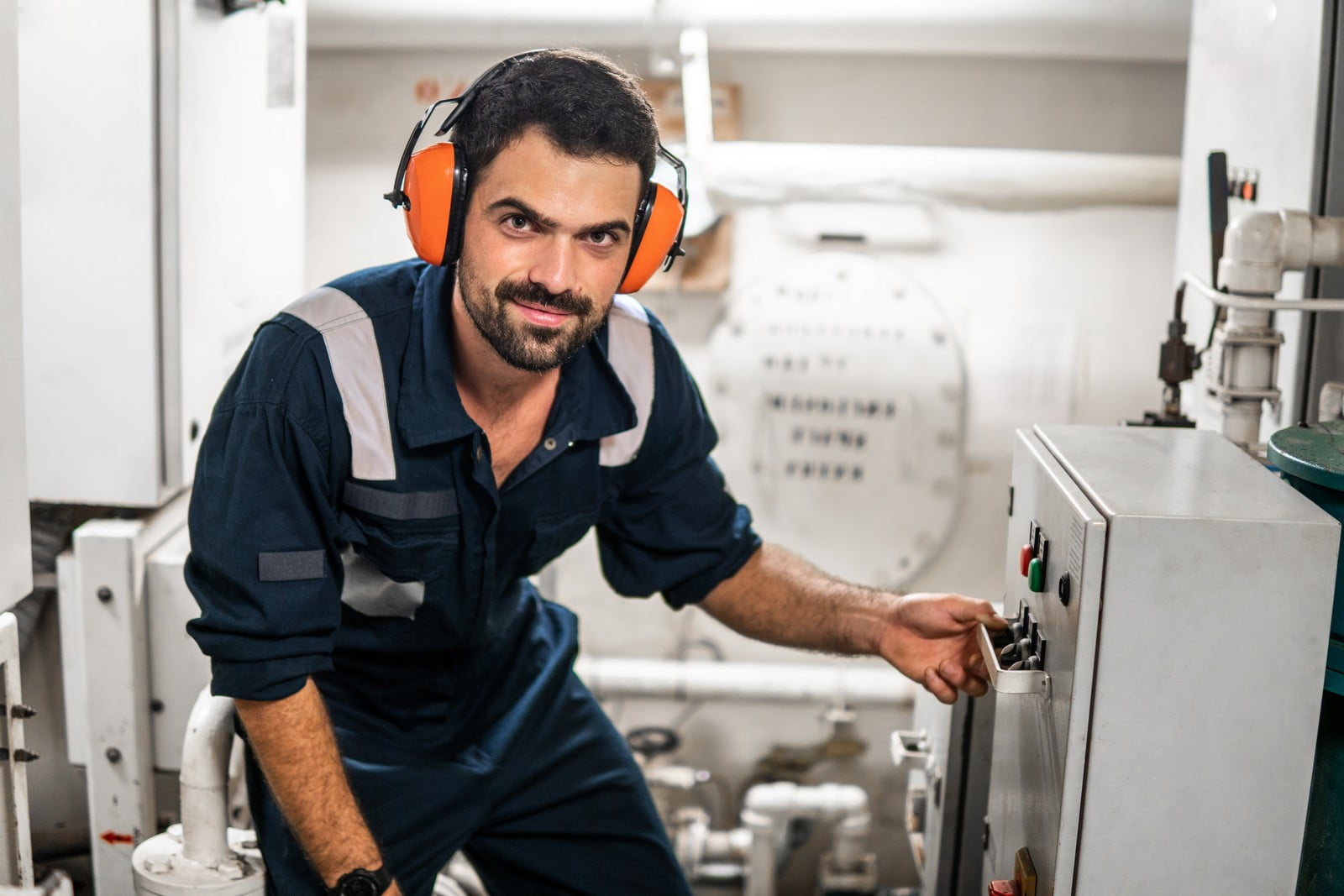 This image features a person who appears to be a worker, technician, or engineer, wearing a dark-coloured work uniform with reflective stripes and protective hearing earmuffs. The individual is squatting next to some kind of machinery or control panel, with his hand on a lever or switch, suggesting he is operating, inspecting, or repairing the equipment. The background shows more industrial equipment and a white panel with various dials and indicators, which indicates that the setting may be some type of workshop, factory, or industrial plant. His expression seems friendly and confident, and he is looking directly at the camera.