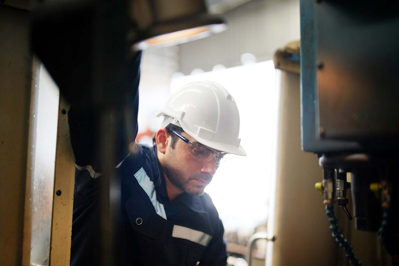 The image shows a person in a work environment wearing a white hard hat, which indicates they might be on a worksite or within an industrial setting where safety precautions are necessary. The individual appears to be focused on inspecting or operating machinery, indicated by their direct gaze and close proximity to what might be equipment or control panels. The person is wearing work attire that includes a dark-coloured shirt with visible cuffs and a high visibility strip, which is common in many industrial and construction workplaces for safety and visibility. The background is blurred, emphasising the subject, but retains the industrial aesthetic with metal structures and possibly machinery.