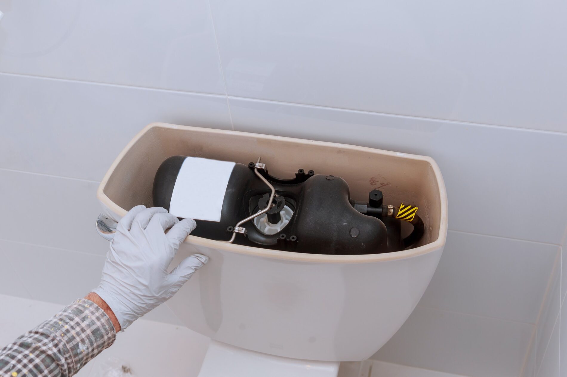 The image shows the inside of a toilet cistern. A person's hand, wearing a white glove, is holding part of the toilet mechanism, specifically the float that is connected to the fill valve. The float is usually responsible for regulating the water level within the cistern. When the float rises to a certain level, it shuts off the water valve to stop the inflow of water, and as the water level drops after a flush, it allows the valve to open and refill the tank. The image shows that someone is either repairing or adjusting the toilet's flushing mechanism. The white and beige colours suggest it may be a residential or commercial grade toilet commonly found in many restrooms.