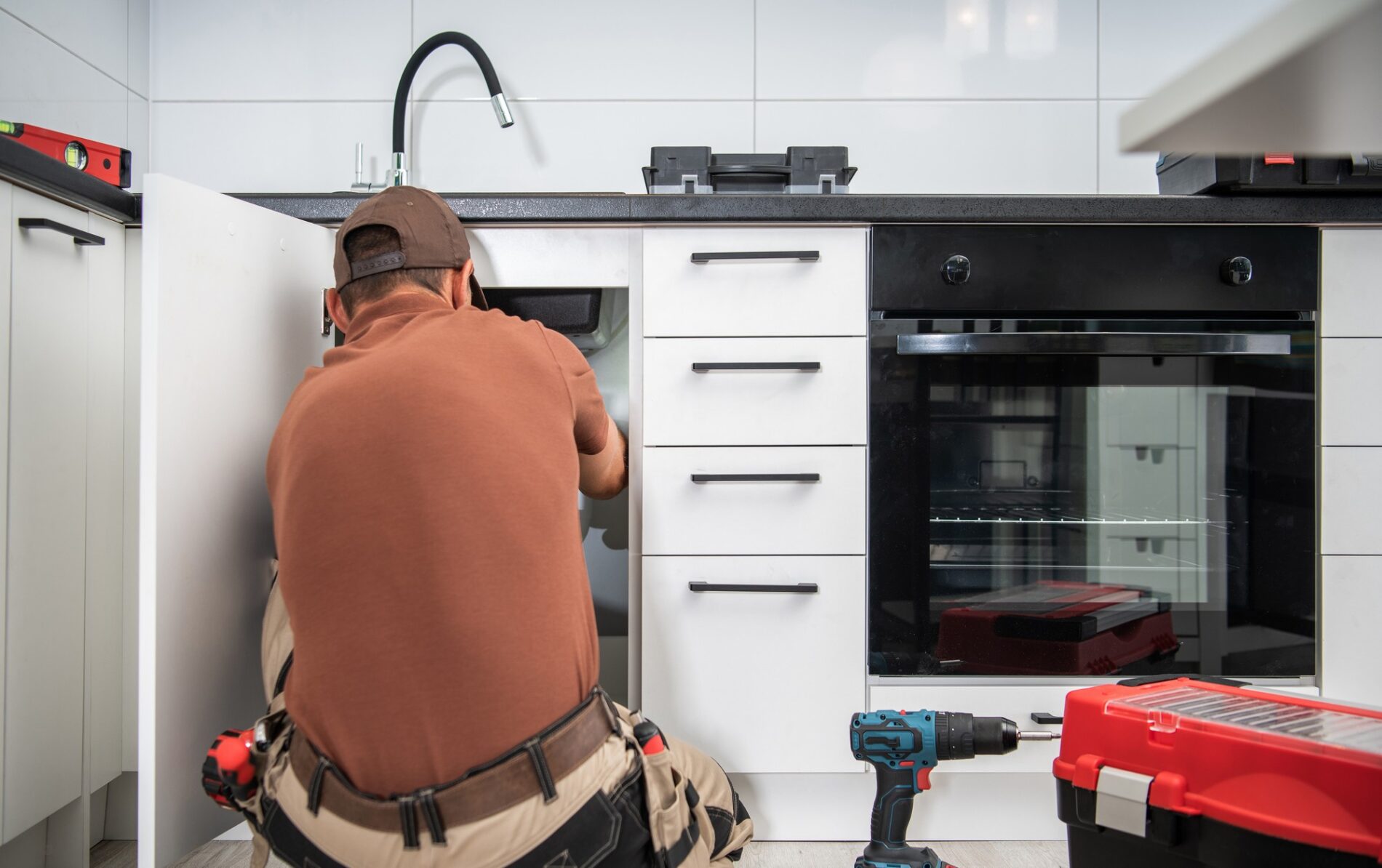 This image shows a person, likely a professional handyman or plumber, working on the plumbing under a kitchen sink. The individual is wearing a cap and a tool belt, indicating they are equipped to perform some sort of maintenance or repair. In front of the person, there's a modern kitchen setup with white cabinets, a black countertop, and built-in appliances like a black oven. To the right of the image, on the countertop, there is a red toolbox and a blue power drill, supporting the idea that some repair or installation work is being carried out. A spirit level is also visible on top of the counter, suggesting the precision required in such work. The kitchen tiles are white, giving the space a clean and sharp appearance.