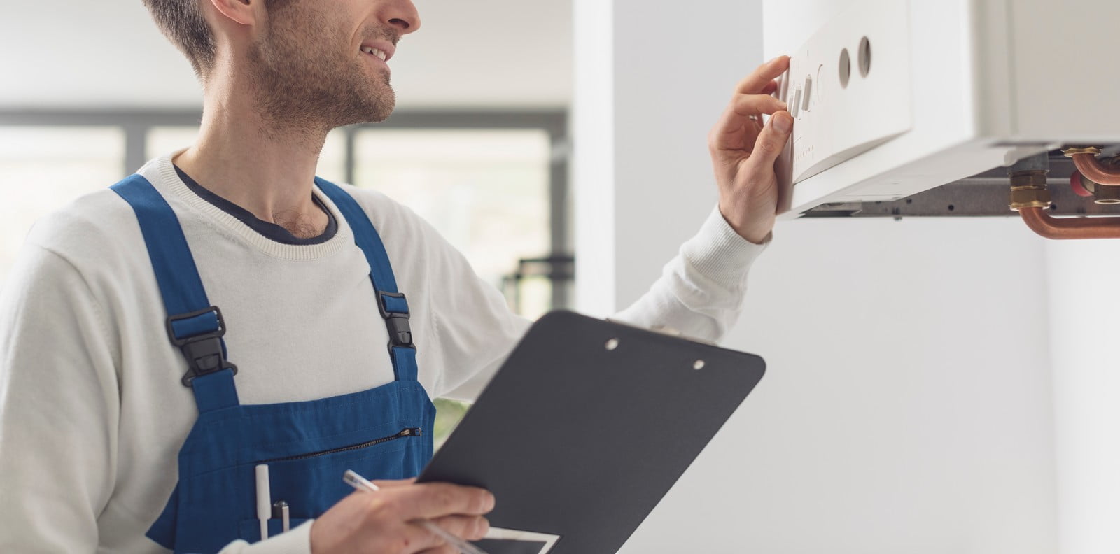 The image shows a person who appears to be a technician or a maintenance worker. Wearing a blue and white work outfit with braces, the individual is adjusting or inspecting a wall-mounted device, which could be a thermostat, control panel, or other type of appliance. The technician is also holding a clipboard, perhaps to record readings or complete a service report. The environment suggests an indoor setting with ample natural light.