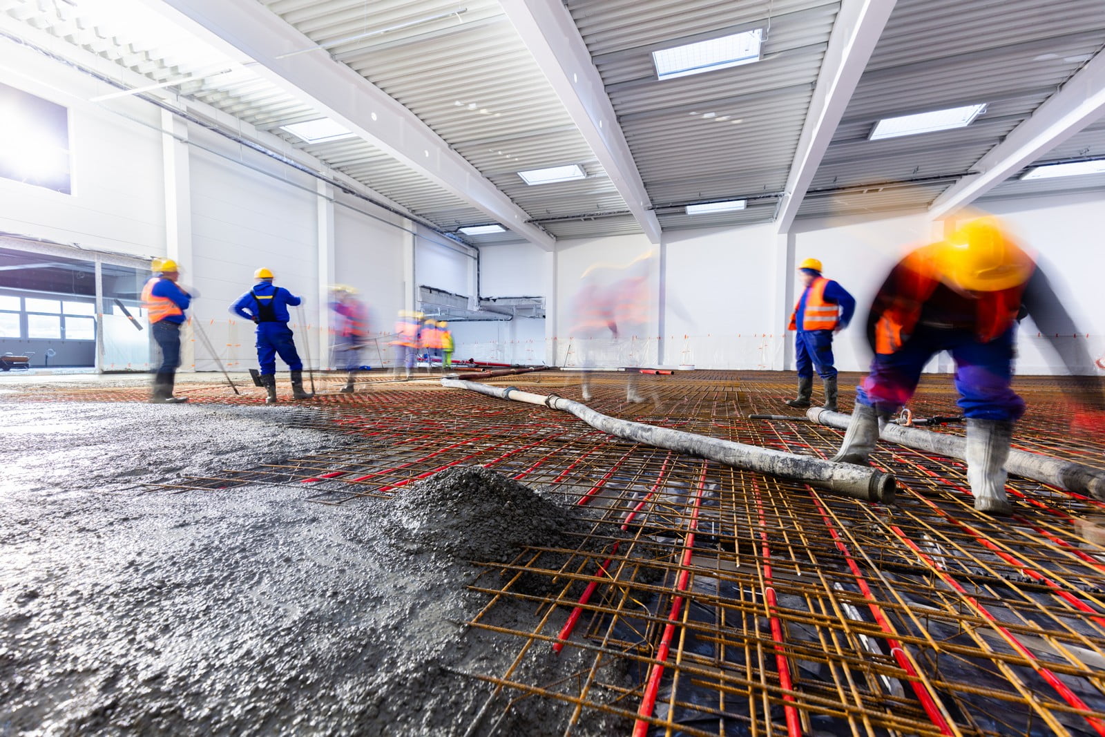 This image shows a construction scene inside what appears to be a large industrial building or warehouse. There are several workers wearing safety helmets and high-visibility vests. The floor is being prepared with a network of steel rebar laid out in a grid pattern, which is a typical reinforcement for poured concrete. A concrete pump hose is visible, and it seems that concrete is being poured and leveled by the workers, which gives the surface a wet and shiny appearance. The blurred motion of the workers indicates activity and movement, often used to convey the action in a photograph. The construction lights and large windows provide illumination for the work area.