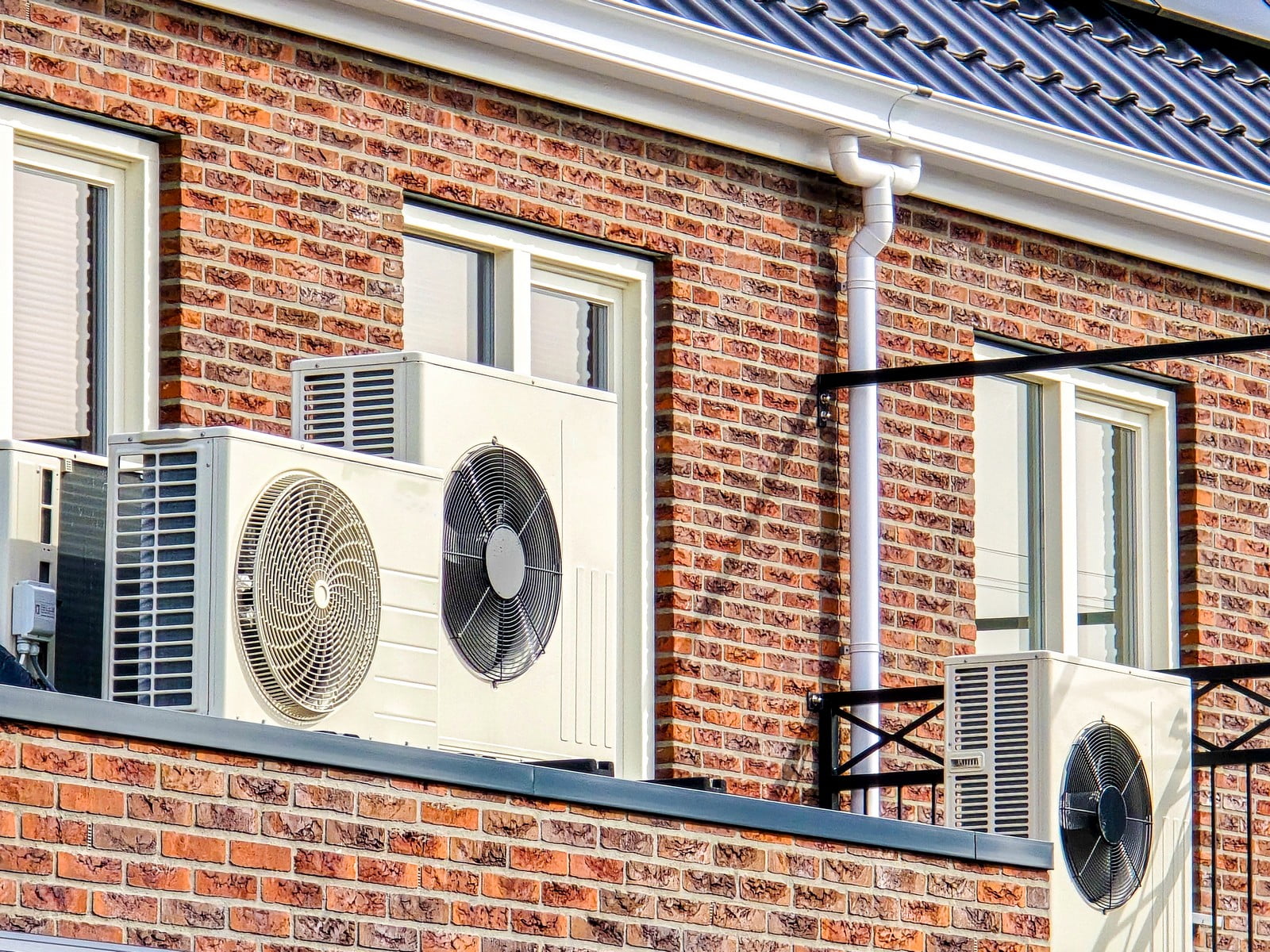 The image shows an exterior wall of a building with three outdoor units of split system air conditioners mounted on brackets. The wall is constructed with red brick, and there are two windows with white frames and closed shutters. Above the wall, there is a portion of the building's roof, which is covered with dark roof tiles. To the right, there is a white pipe, presumably part of the building's plumbing or ventilation system, running vertically along the wall and then turning at an angle towards the roof. The time appears to be during the day with sunlight illuminating the building's facade.