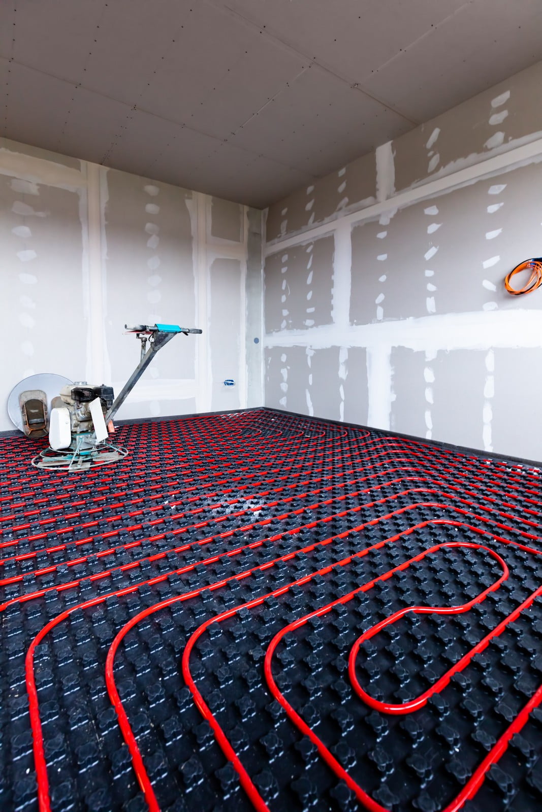 The image shows an underfloor heating installation in progress. You can see the red pipes arranged in loops and laid out across the floor, which will carry hot water to heat the room from below. The floor is covered with black insulation panels which are likely designed to ensure efficient heat distribution and minimise heat loss. Additionally, a concrete screed needs to be poured over these pipes to encase them before the final flooring is installed.On the side, there is a tool, possibly used for the installation of the heating system. The background shows the walls in an unfinished state, with marks suggesting that they have been recently drywalled or plastered, supported by the presence of joint tape and patches of joint compound that have not yet been sanded smooth or painted. An electrical cable reel can also be observed on the right side. The overall scene suggests an environment under renovation or construction.