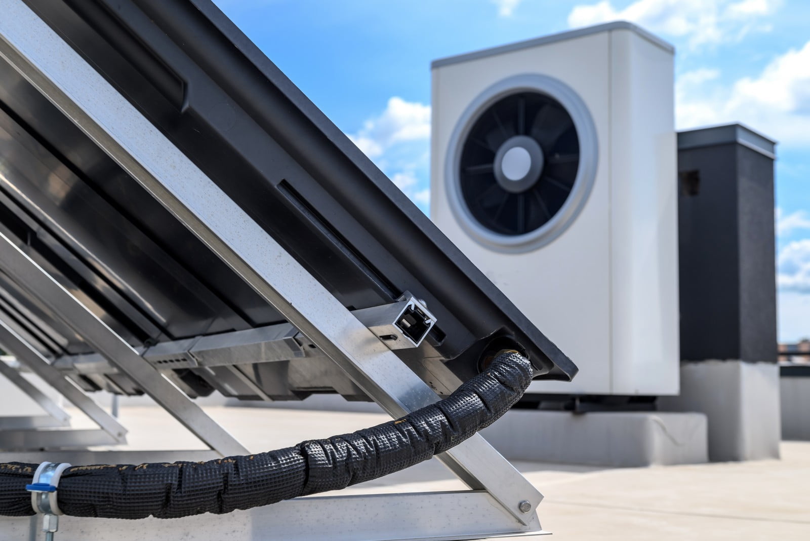 This image shows a rooftop with solar panels and a heating ventilation and air conditioning (HVAC) unit. The foreground features a closer view of the side of a solar panel angled to capture sunlight, including the mounting hardware and a conduit with cables, likely for carrying the electrical current generated by the panels. In the background, there is a large HVAC unit, which is used to regulate the temperature within a building. This setup indicates a combination of renewable energy utilization through the solar panels and climate control systems on the roof of a modern building.