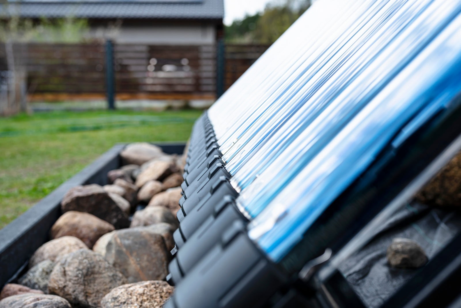 The image shows a close-up, angled view of a structure that appears to be an outdoor feature, likely part of a building or a standalone structure. The focus is on elongated, translucent blue panels that are arranged in a row and supported by a black framework. The panels are likely made of a lightweight, synthetic material, possibly polycarbonate or acrylic, and could be serving as a roof or canopy—perhaps for a greenhouse, patio cover, or a similar application.Below the blue panels, there is a glimpse of what appears to be a garden or yard area. You can see the corner of a black, raised garden bed or planter box filled with large, smooth rocks or river stones, indicating an element of landscape design.The background is out of focus but suggests a residential setting with a lush green lawn and a wooden fence. The overcast sky suggests it might be a cloudy day. The perspective and selective focus of the image emphasize the materials and construction details of the panels and the framework, drawing attention to the texture and colour contrasts.