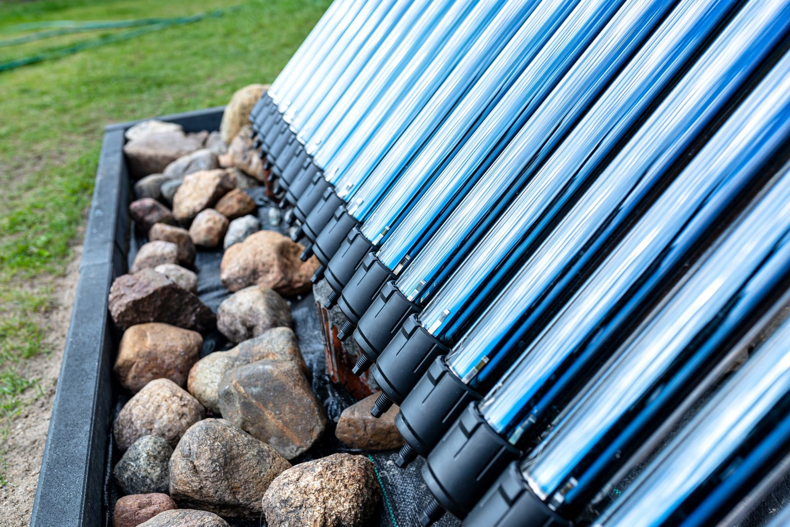 This image features a collection of solar vacuum tubes as part of a solar water heating system. These tubes are typically made of glass and use the greenhouse effect to capture heat from the sun, which is then used to heat water inside the system. The background shows a grassy area, and the solar tubes are installed on a frame that includes a bed of rocks—likely for both support and aesthetic purposes. The solar water heating system appears to be outdoor, suggesting that it is part of a residential or commercial building's renewable energy installation.