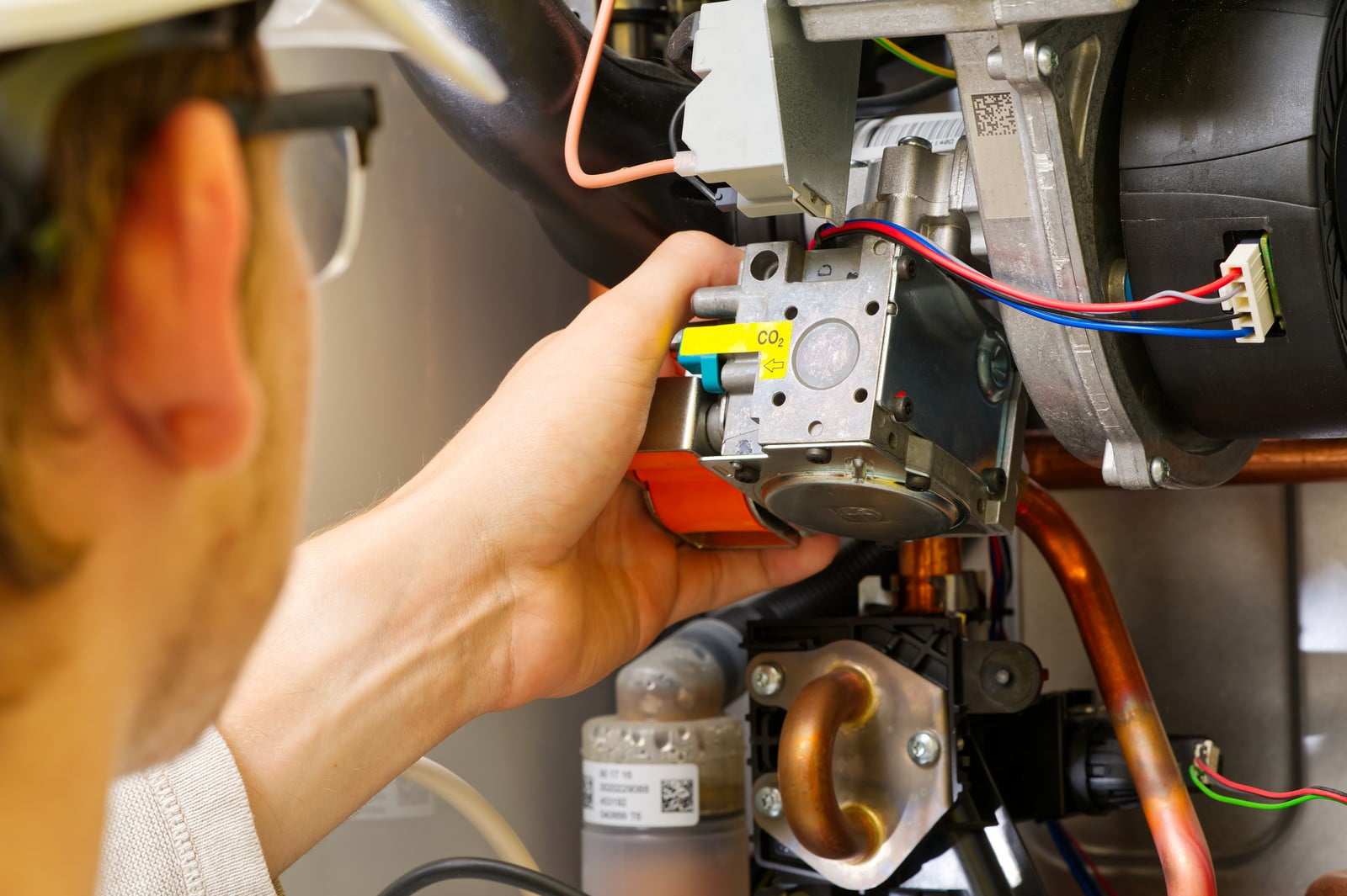 This image shows a person working on what appears to be a component of an HVAC (heating, ventilation, and air conditioning) system, specifically a part of a furnace or a boiler. The individual, only partially visible, is wearing a safety helmet, which suggests they are a professional technician or engineer. The person is handling a motor or pump, which is part of the mechanical system. The yellow label on the component with "CO2" suggests that they might be adjusting or inspecting the petrol levels or combustion process. There are copper pipes and electrical wirings visible as well, indicating a complex mechanical setup.