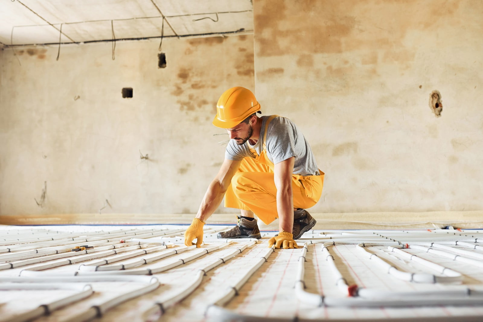 This image shows a construction worker, dressed in a safety helmet and gloves, working on the installation of an underfloor heating system. He is in a crouched position, attending to the network of pipes laid out across the floor, which are part of the heating system configuration. The environment appears to be an unfinished room undergoing renovation or construction, with an incomplete ceiling and bare walls, emphasising the under-construction status.