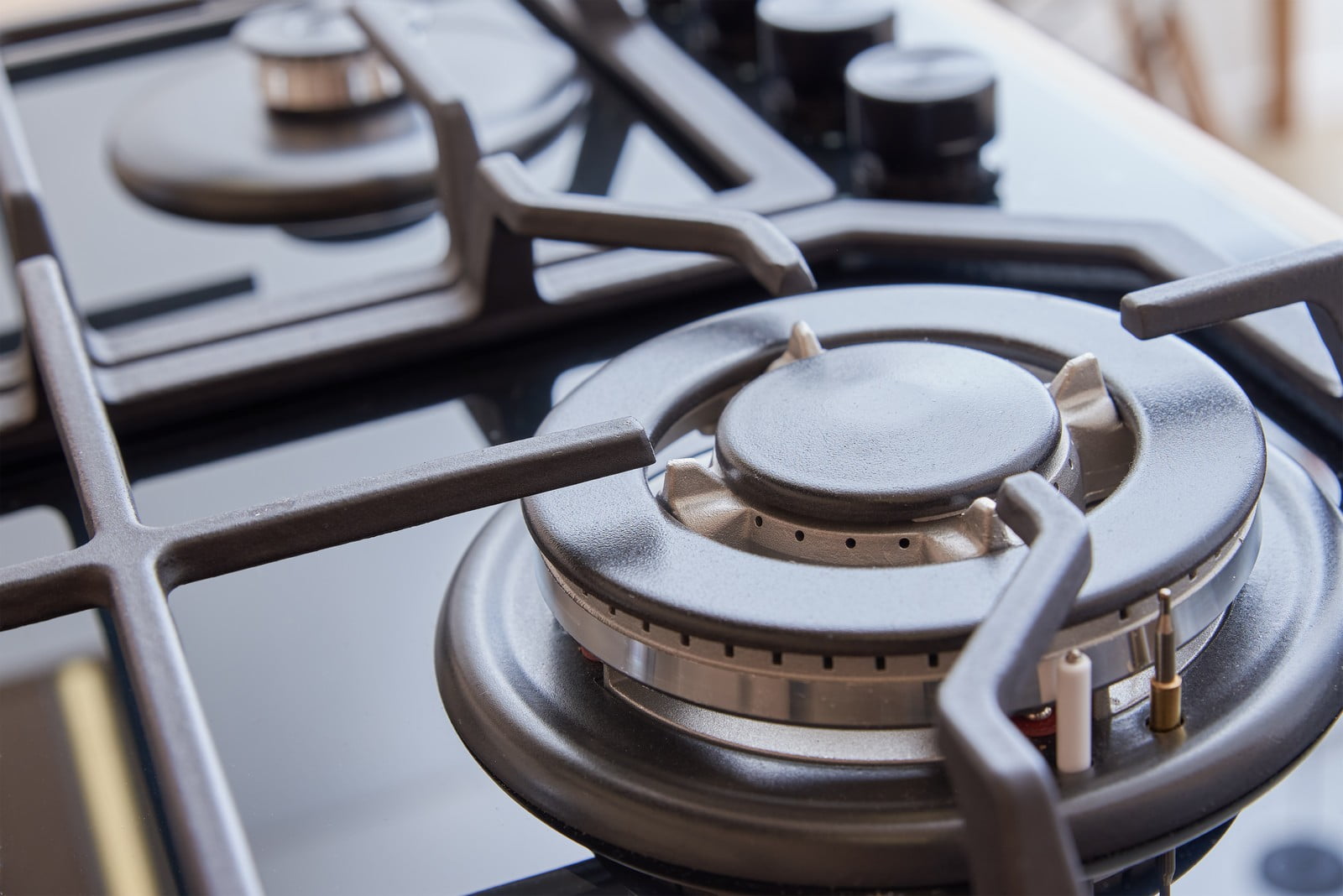 The image showcases a modern, stainless steel petrol stove with four burners, neatly installed in a contemporary kitchen setting. Each burner is topped with a sleek, cast-iron grate that provides a stable surface for pots and pans. The stovetop features an array of control knobs located at the front, each with clear labelling for precise flame adjustment. Surrounding the stovetop is a smooth, light-coloured countertop, accentuating the clean lines and minimalist design of the kitchen. The background reveals a segment of the kitchen backsplash, made of light, glossy tiles that reflect the ambient lighting, further enhancing the kitchen's bright and airy atmosphere. The overall scene conveys a sense of order and modern functionality, ideal for a cooking enthusiast.