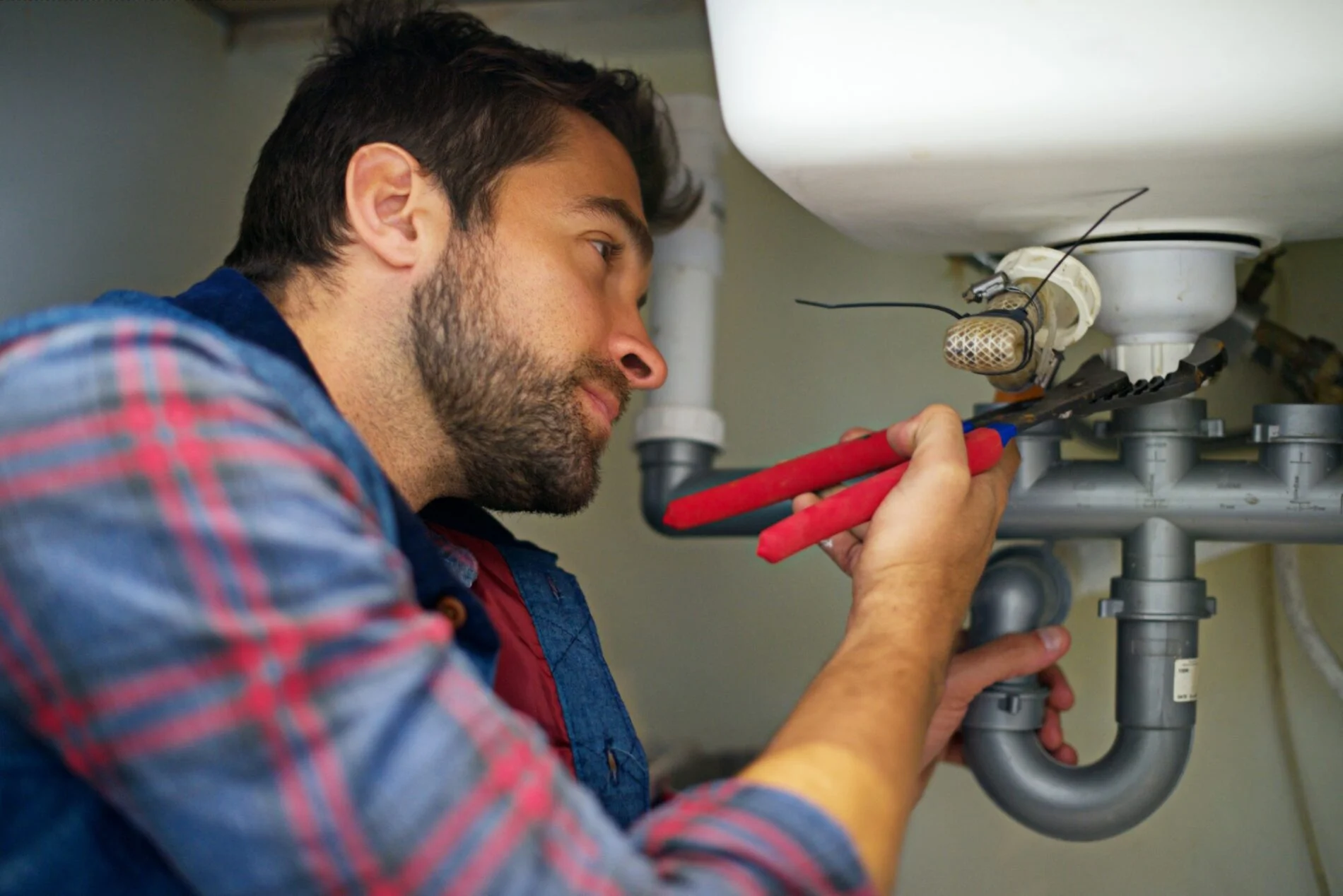 The image shows a professional plumber engaged in fixing a plumbing issue in a well-lit indoor setting. The plumber, a middle-aged man with short dark hair, is dressed in a navy-blue work uniform with a company logo on the chest pocket. He is kneeling on the tiled floor, focused intently on his task. The scene captures him mid-repair, with his toolbox open beside him, displaying various tools and equipment.He is holding a spanner with both hands as he works on tightening a pipe under a sink. The surroundings are clean and organized; the under-sink cabinet is open, revealing plumbing pipes and some cleaning supplies stored within. The sink and countertop are modern, perhaps in a residential kitchen or bathroom. Bright overhead lights illuminate the workspace, highlighting the plumber's meticulous attention to detail as he ensures the connections are secure. His expression is one of concentration and professionalism, reflecting his expertise in the field.